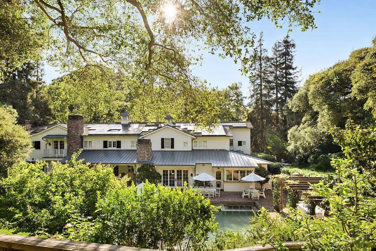 This rear view of a large, multi-story residence showcases a classic farmhouse-inspired aesthetic with white siding, a dark metal roof, and multiple brick chimneys. The home features a spacious brick patio area equipped with outdoor dining furniture and umbrellas, overlooking a swimming pool nestled in a lush, tree-filled backyard. The perspective is elevated, capturing the serene integration of the home's architecture with the surrounding mature trees and natural landscape.