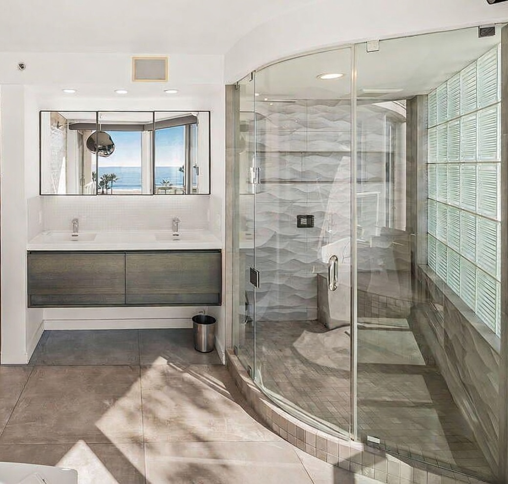 This modern primary bathroom features a floating dual-sink vanity with dark wood-toned cabinetry and a large mirror reflecting a scenic ocean view. To the right, a curved glass-enclosed shower stall showcases textured, wavy-patterned wall tiles and a wall of glass blocks that allow natural light to filter through. The space is finished with large-format neutral floor tiles, creating a clean, contemporary, and airy atmosphere.