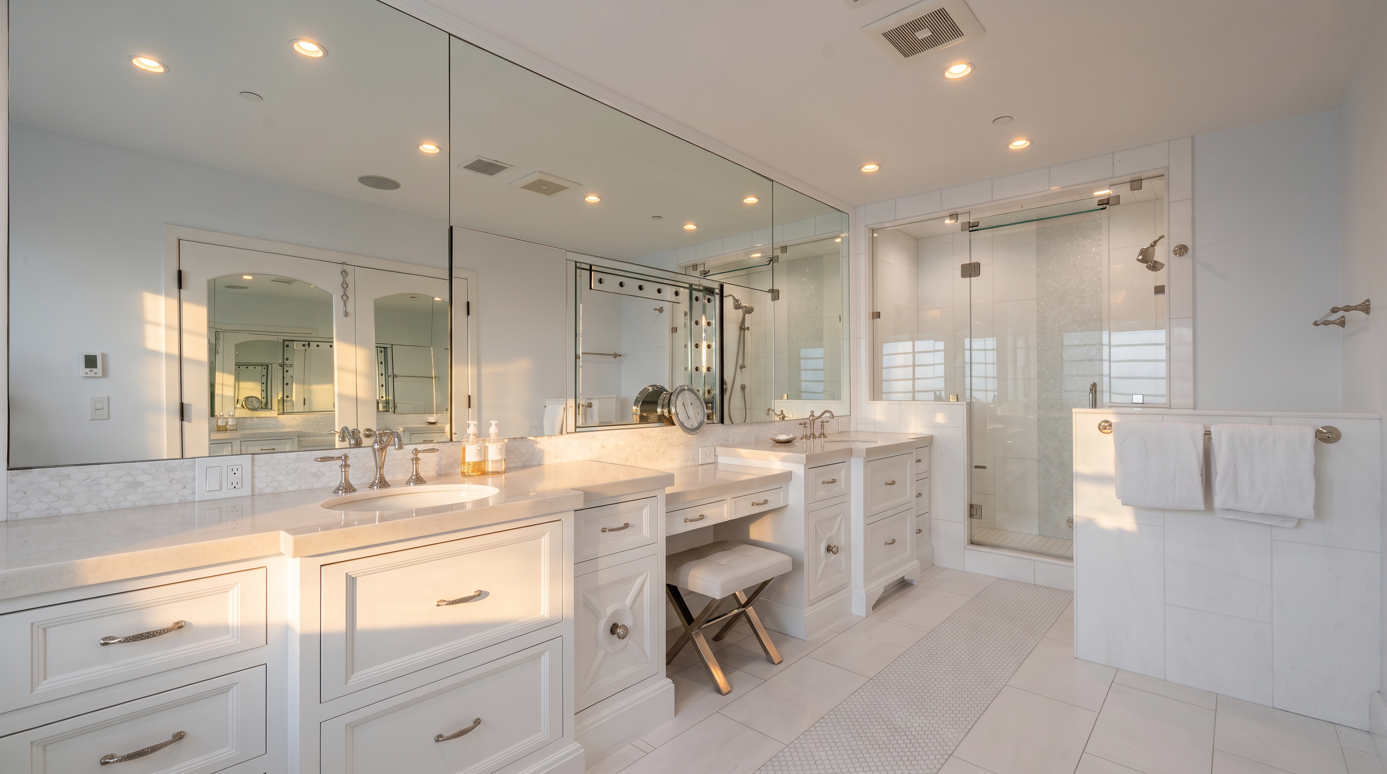 This bright and spacious primary bathroom features a long, white double vanity with stone countertops and a central makeup vanity area. A large, wall-to-wall mirror reflects the room, while a glass-enclosed shower stall sits to the right, adjacent to a tiled wall with towel racks. The space is finished with light-colored tile flooring and recessed lighting, creating a clean and airy atmosphere.