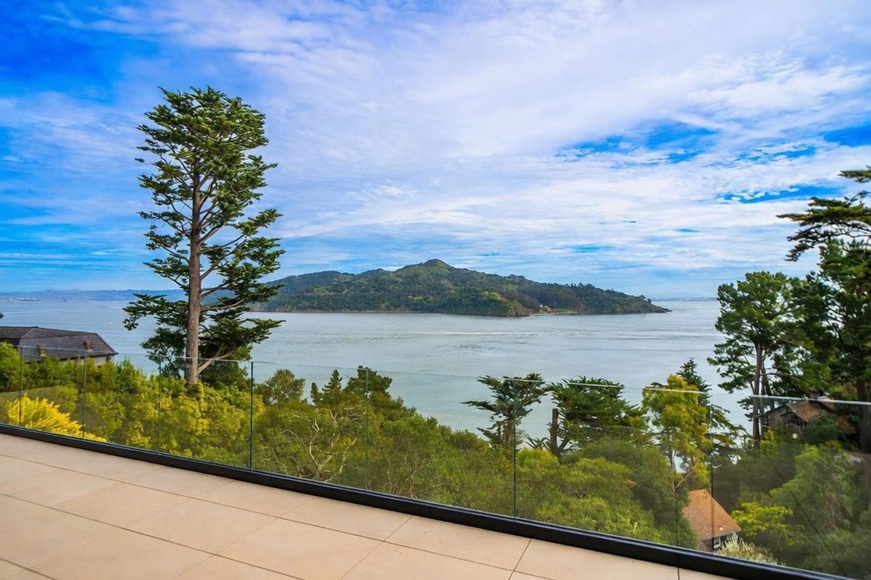 This image captures a stunning, elevated balcony view overlooking a wide body of water with a prominent island in the distance. The balcony features light-toned stone flooring and a modern glass railing that provides an unobstructed, panoramic perspective of the bay and the passing ferry. Lush green trees frame the sides of the view, creating a serene and private atmosphere that emphasizes the property's prime waterfront location.