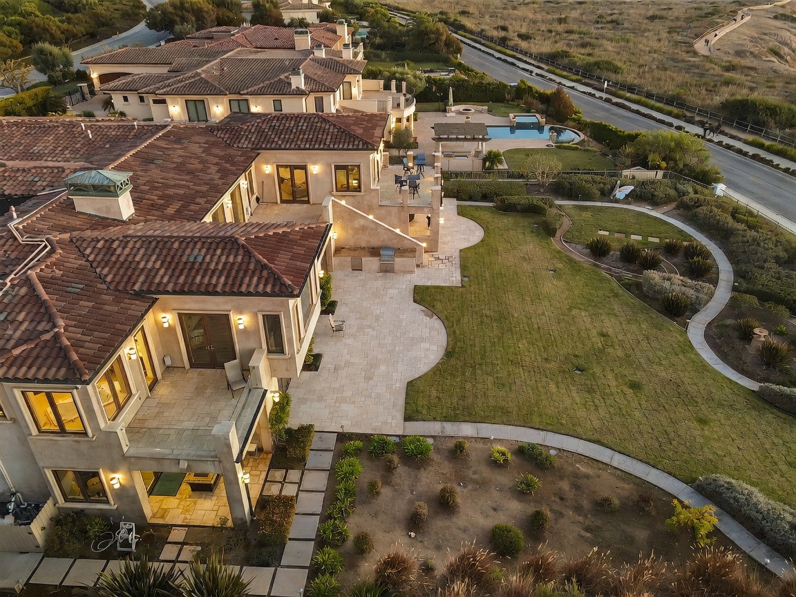 This high-angle aerial view captures a sprawling Mediterranean-style estate with a multi-level tiled roof and extensive outdoor living spaces. The property features a large stone-paved patio, a manicured lawn, and a separate pool area with a spa, all set against a backdrop of rolling hills and a nearby road. The architecture showcases classic design elements, including arched doorways and multiple terraces that overlook the expansive backyard.