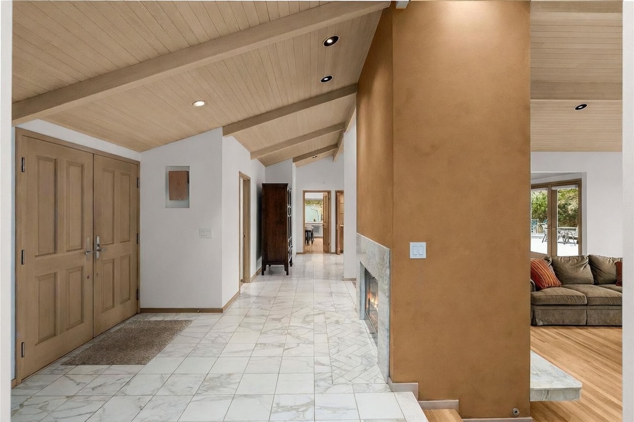 This entryway and hallway area features light-colored patterned tile flooring and a vaulted ceiling with exposed beams. A prominent, warm-toned wall separates the entry from the living space, incorporating a fireplace with a stone surround. The perspective looks down the main corridor, showcasing a bright, open layout that leads toward additional rooms and a glimpse of the living area to the right.