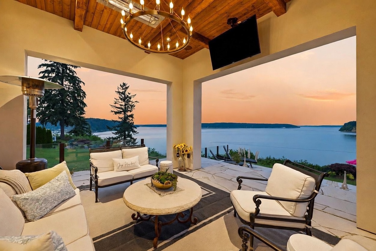 This covered outdoor living space offers a sophisticated lounge area with comfortable seating, a central stone-topped coffee table, and a large chandelier hanging from a wood-paneled ceiling. The space opens up to a breathtaking, unobstructed view of a calm body of water at sunset, framed by large structural pillars. The setting is serene and luxurious, perfect for evening relaxation while overlooking the expansive waterfront landscape.