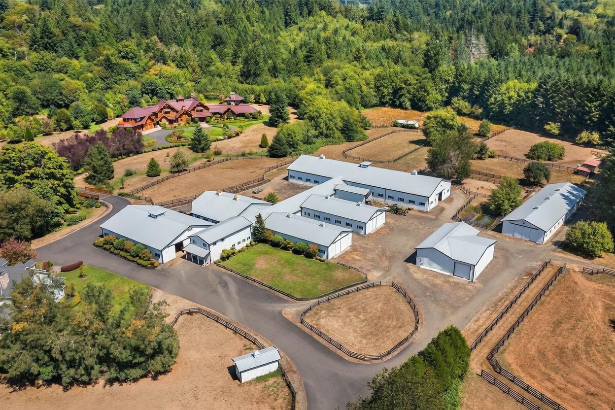 This expansive aerial view captures a sprawling equestrian-style estate nestled within a lush, forested landscape. The property features a large, multi-gabled main residence set back from a complex of white-sided outbuildings, barns, and storage structures connected by paved driveways. The grounds are divided into multiple fenced paddocks and open fields, showcasing a serene and well-maintained rural setting.