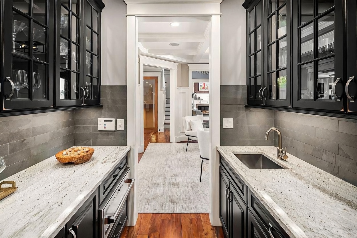 This elegant butler's pantry features dark cabinetry paired with light, patterned stone countertops and a sleek undermount sink. A gray tile backsplash adds a sophisticated texture, while glass-front upper cabinets provide a refined display space. The perspective looks through the pantry into a bright, open living area, highlighting the home's seamless flow and high-end finishes.