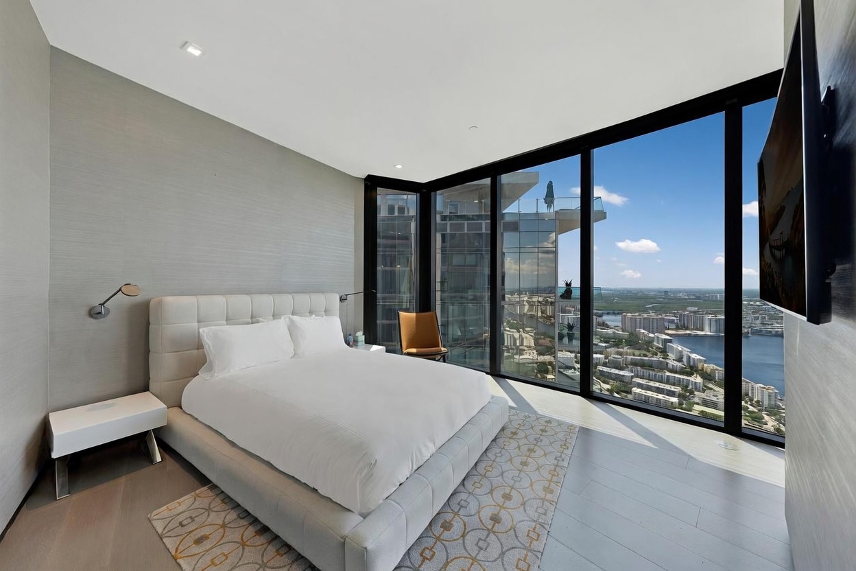 This modern primary bedroom features a plush, upholstered bed centered against a textured gray wall, flanked by minimalist white nightstands. The room is defined by expansive floor-to-ceiling windows that provide a panoramic, high-altitude view of the city skyline and waterfront. A patterned area rug adds a touch of geometric interest to the light-toned flooring, while a single accent chair sits near the glass, creating a serene and sophisticated atmosphere.