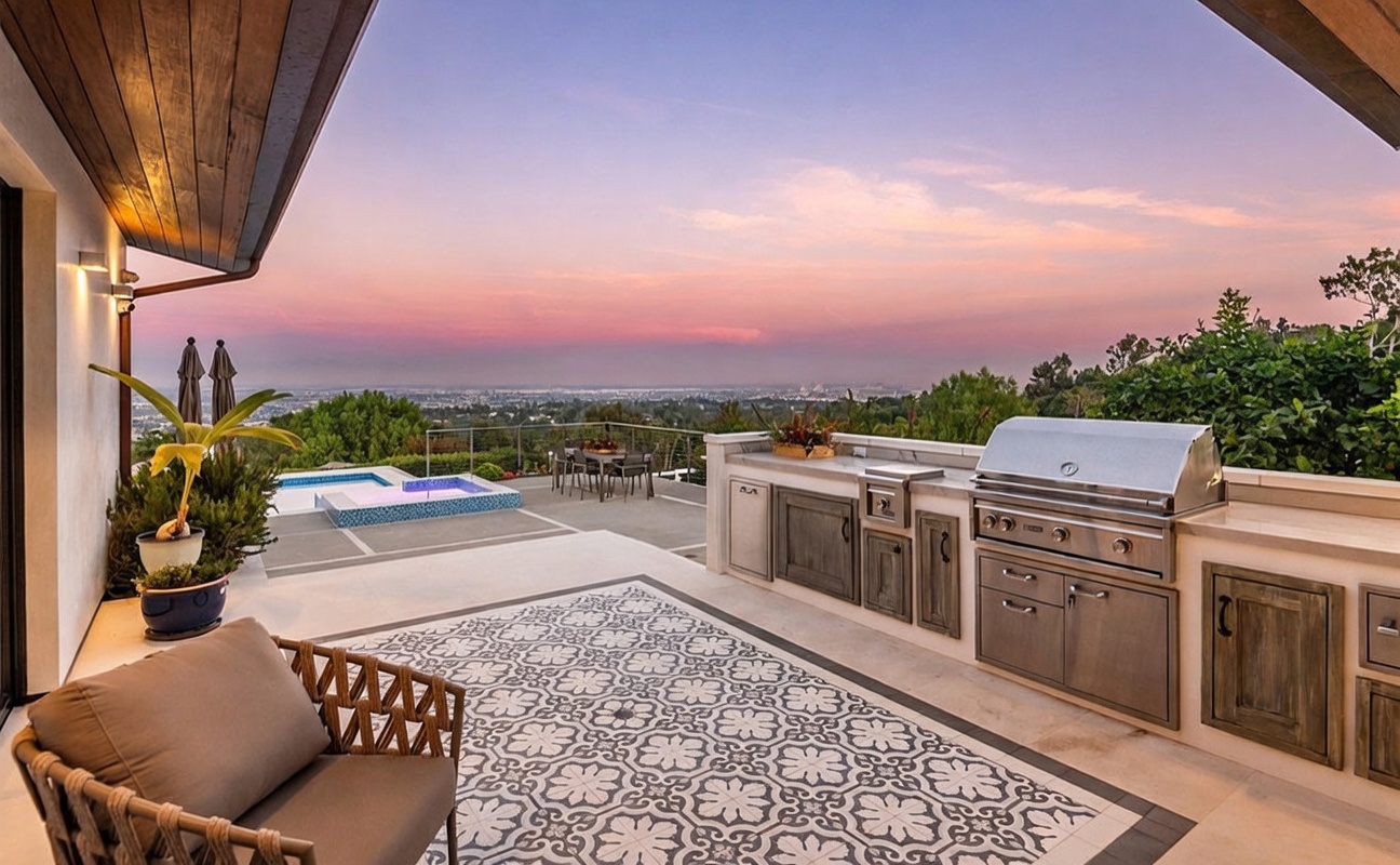 This expansive outdoor living space features a covered patio area equipped with a built-in stainless steel grill and cabinetry, perfect for al fresco dining. The space transitions seamlessly to an open deck area that includes a pool and spa, all set against a breathtaking panoramic view of the city skyline at sunset. The area is furnished with a comfortable lounge chair and a patterned outdoor rug, creating a sophisticated and inviting atmosphere for relaxation or entertaining.
