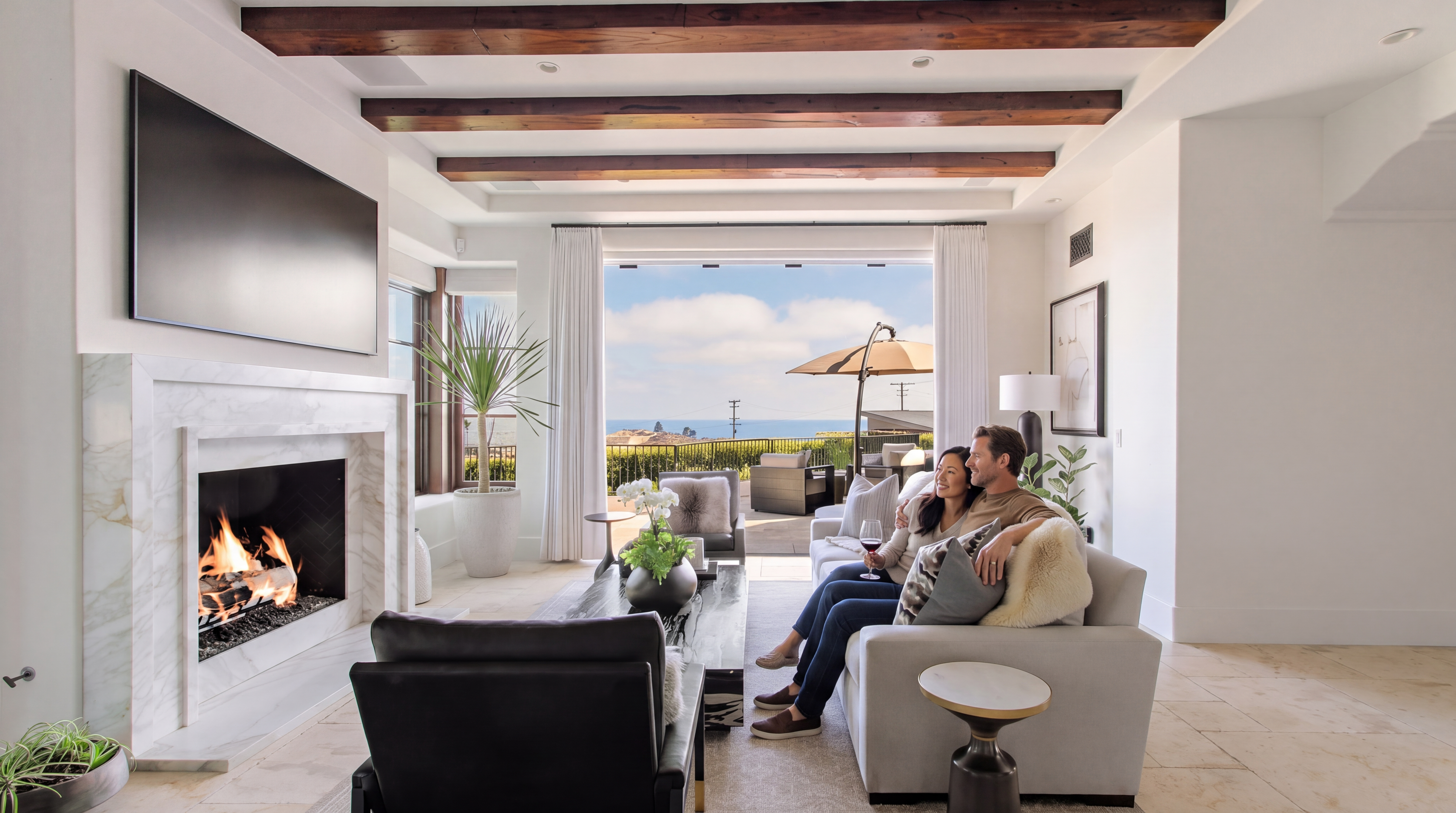 This elegant and inviting living room features a beautiful marble fireplace as its focal point, complemented by exposed wood ceiling beams that add a rustic touch. The open-concept design seamlessly connects the indoor living space to a stunning patio with an ocean view through large sliding glass doors. The room is tastefully furnished with comfortable seating, creating a warm and luxurious atmosphere perfect for relaxation.
