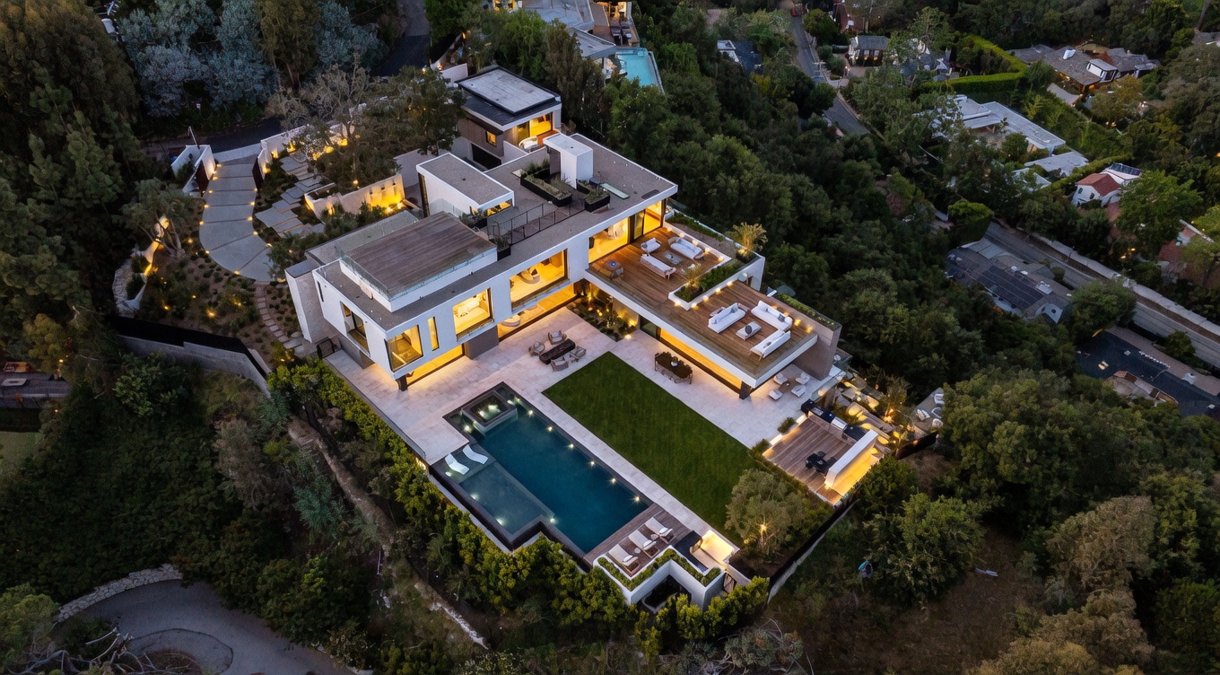 This stunning aerial perspective captures a contemporary multi-level luxury residence nestled into a lush, wooded hillside. The property features a sprawling rectangular swimming pool, a manicured lawn, and expansive tiered wooden decks furnished for outdoor living. The architecture emphasizes clean lines and large glass panels, creating a seamless transition between the sophisticated interior spaces and the surrounding natural landscape.