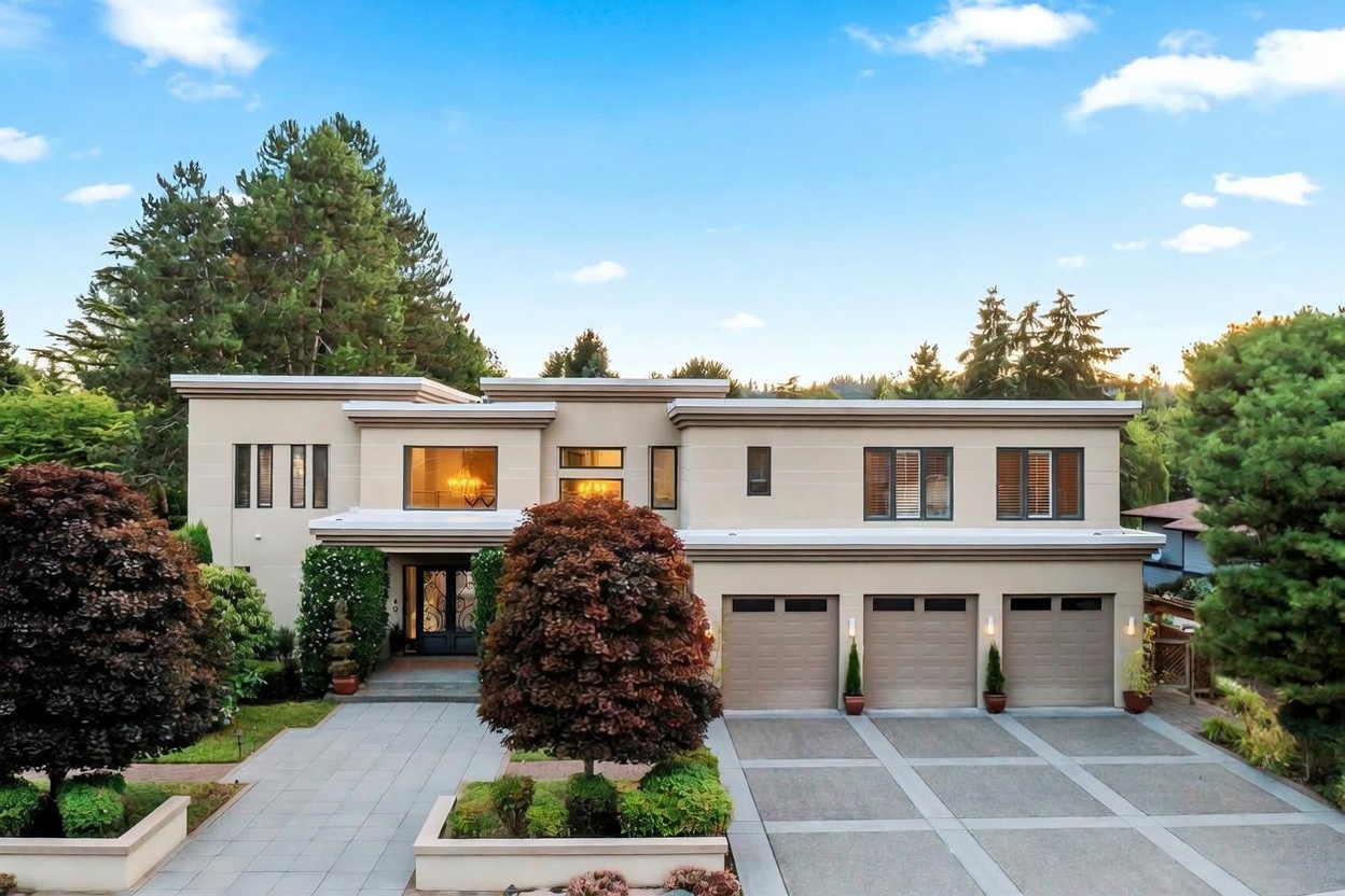 This modern, two-story residence features a clean, light-toned facade with a symmetrical design and a three-car garage. The front entrance is accessed via a paved walkway flanked by manicured landscaping, including a prominent ornamental tree in the foreground. The property is set against a backdrop of mature evergreen trees under a clear blue sky, presenting a sophisticated and well-maintained curb appeal.