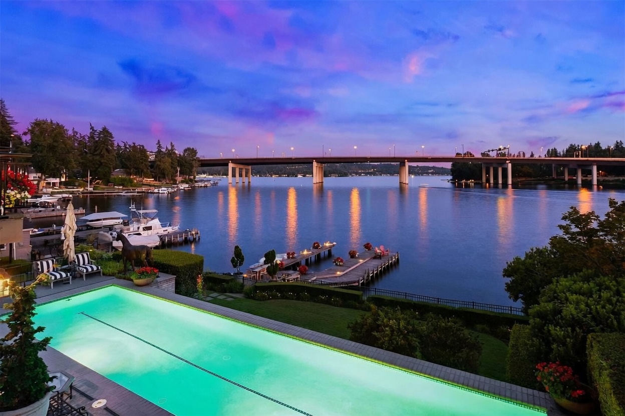 This stunning twilight view captures a luxurious rectangular swimming pool in the foreground, overlooking a serene lake and a distant bridge. The scene features a private wooden dock extending into the water, accented by warm lighting, while the sky transitions into a vibrant purple and blue sunset. The perspective is elevated, providing a cinematic vantage point that emphasizes the property's waterfront lifestyle and tranquil atmosphere.