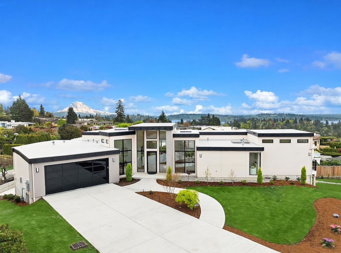 This modern, single-story residence features a striking contemporary design with clean lines, a flat roof, and a prominent glass-enclosed entryway. The exterior is finished in light-toned siding, complemented by a large black garage door and expansive windows that create a bright, open aesthetic. Set against a backdrop of a clear blue sky and a distant mountain peak, the property includes a manicured lawn and a curved concrete driveway leading to the entrance.