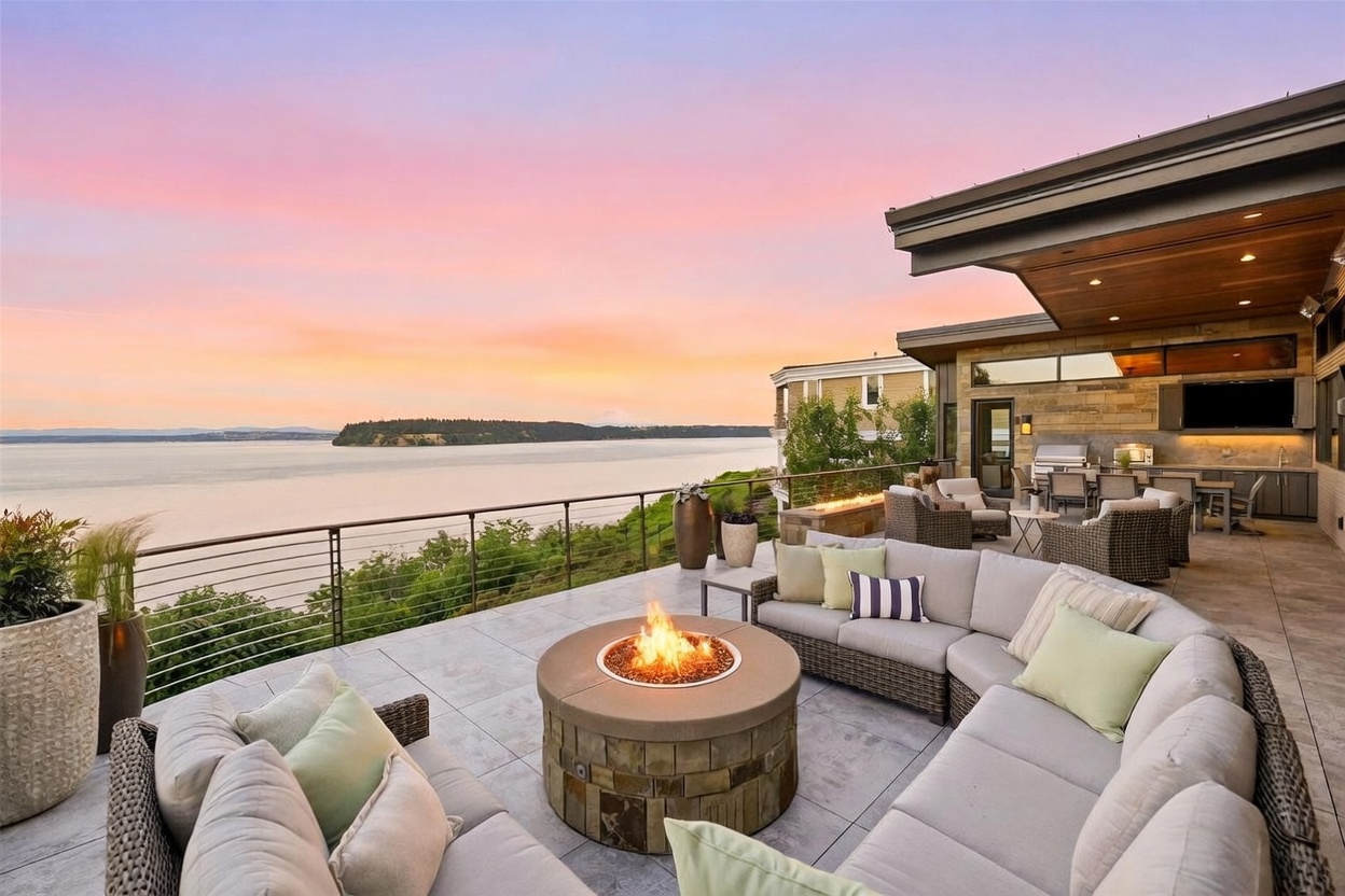 This expansive, modern outdoor terrace offers a panoramic view of the water and distant mountains under a vibrant sunset sky. The space is furnished with a large, curved sectional sofa arranged around a circular stone fire pit, creating an inviting lounge area. Further back, an outdoor kitchen and dining setup are positioned under a covered section of the deck, making this an ideal space for entertaining.