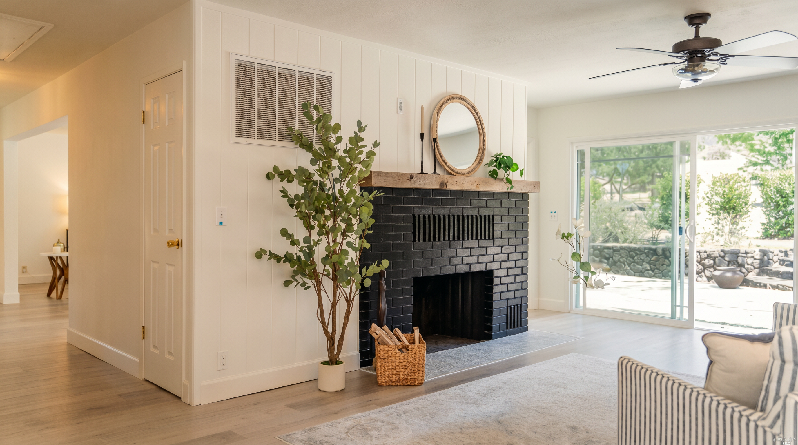 This bright living room features a striking black brick fireplace as its focal point, topped with a natural wood mantel and a decorative mirror. The space is anchored by a large, round woven ottoman and a light-toned area rug, set against white vertical paneling and light wood-look flooring. A sliding glass door provides a seamless transition to the outdoor patio, filling the room with natural light.