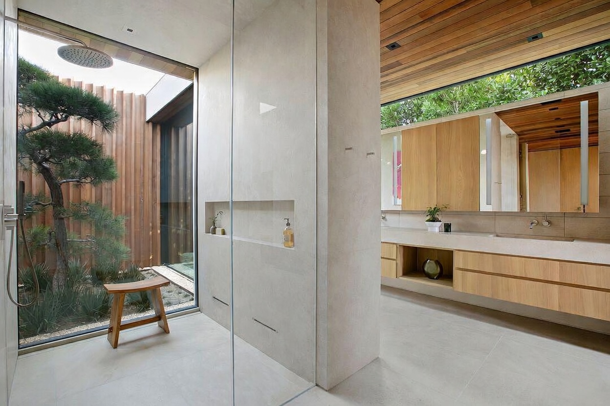 This luxurious, modern primary bathroom features a seamless glass-enclosed walk-in shower that opens directly to a private, zen-inspired outdoor garden area. The space is characterized by high-end finishes, including light wood cabinetry, a floating vanity, and a striking wood-slat ceiling that adds warmth to the minimalist design. The perspective captures the harmonious blend of indoor and outdoor living, emphasizing the serene, spa-like atmosphere of the home.