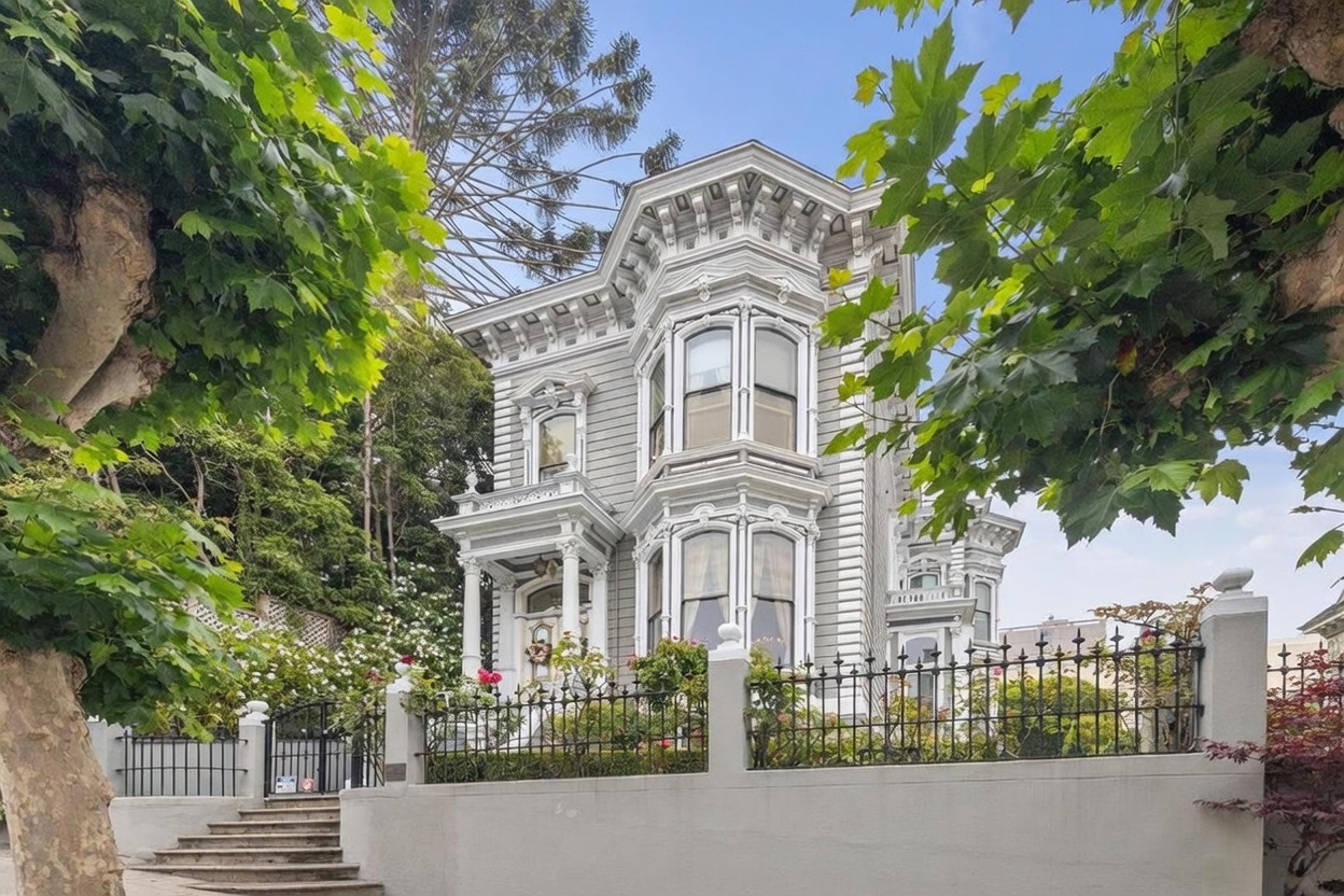 This striking Victorian-style home features intricate architectural detailing, including ornate cornices, bay windows, and light gray horizontal siding. The property is elevated above the street level, accessed by a stone staircase, and enclosed by a decorative black iron fence atop a solid concrete wall. Lush green trees frame the house, creating a classic and elegant curb appeal typical of historic urban architecture.