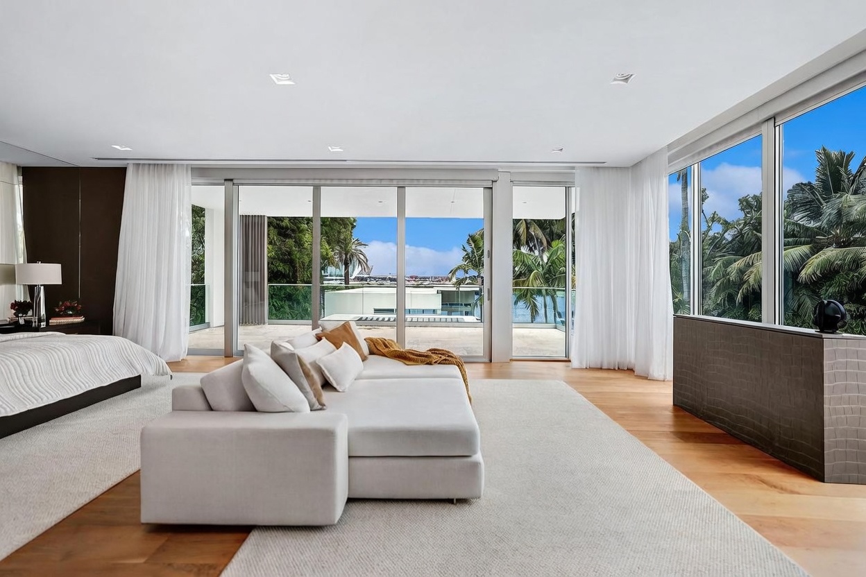 This spacious primary bedroom features a neutral color palette and a large, plush sectional sofa positioned in the center of the room. The space is defined by expansive floor-to-ceiling glass sliding doors that open onto a private balcony, offering panoramic views of the surrounding greenery and water. Light wood flooring and minimalist decor create a serene, high-end atmosphere, while a textured wall panel adds a touch of modern sophistication.