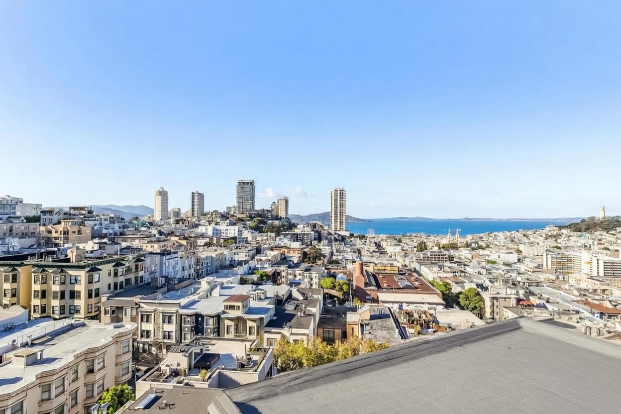 This high-angle aerial view captures a sprawling urban cityscape, showcasing a dense collection of residential buildings and rooftops under a clear blue sky. In the distance, the iconic San Francisco skyline is visible, framed by the sparkling waters of the bay. The perspective provides a comprehensive look at the neighborhood's density and its proximity to the waterfront, emphasizing the expansive city views.