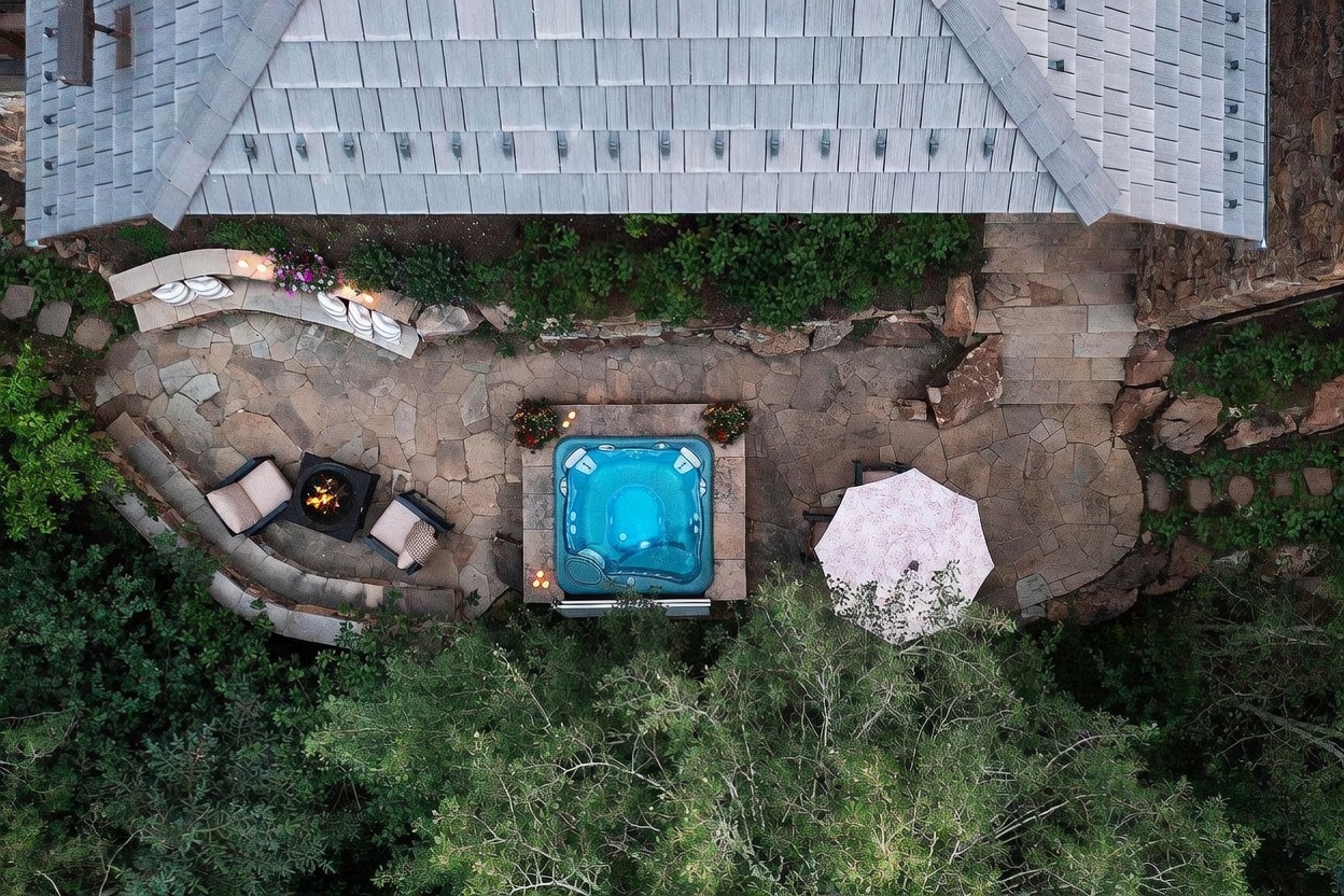 This aerial perspective captures a secluded stone patio nestled against a home with a gray shingled roof. The outdoor living space features a central hot tub, a fire pit area with curved stone seating, and a dining spot shaded by a patterned umbrella. Surrounded by lush greenery and natural rock landscaping, the area offers a private and tranquil retreat.