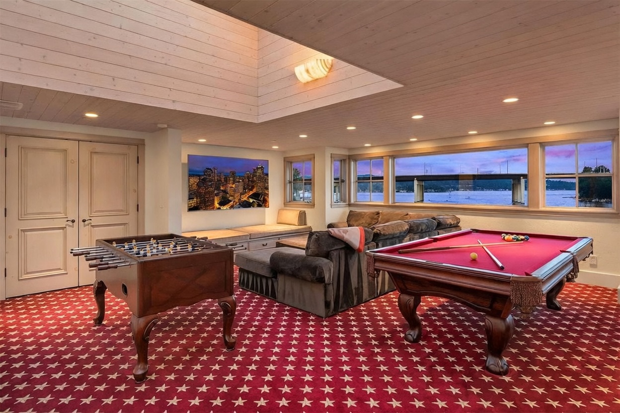 This spacious recreation room features a vibrant red carpet with a repeating star pattern, creating a playful and classic game room atmosphere. The space is equipped with a foosball table, a pool table, and a comfortable sectional sofa positioned in front of a wall-mounted television. Large windows provide a scenic view of the water and a bridge, while a recessed ceiling area with integrated lighting adds architectural interest to the room.