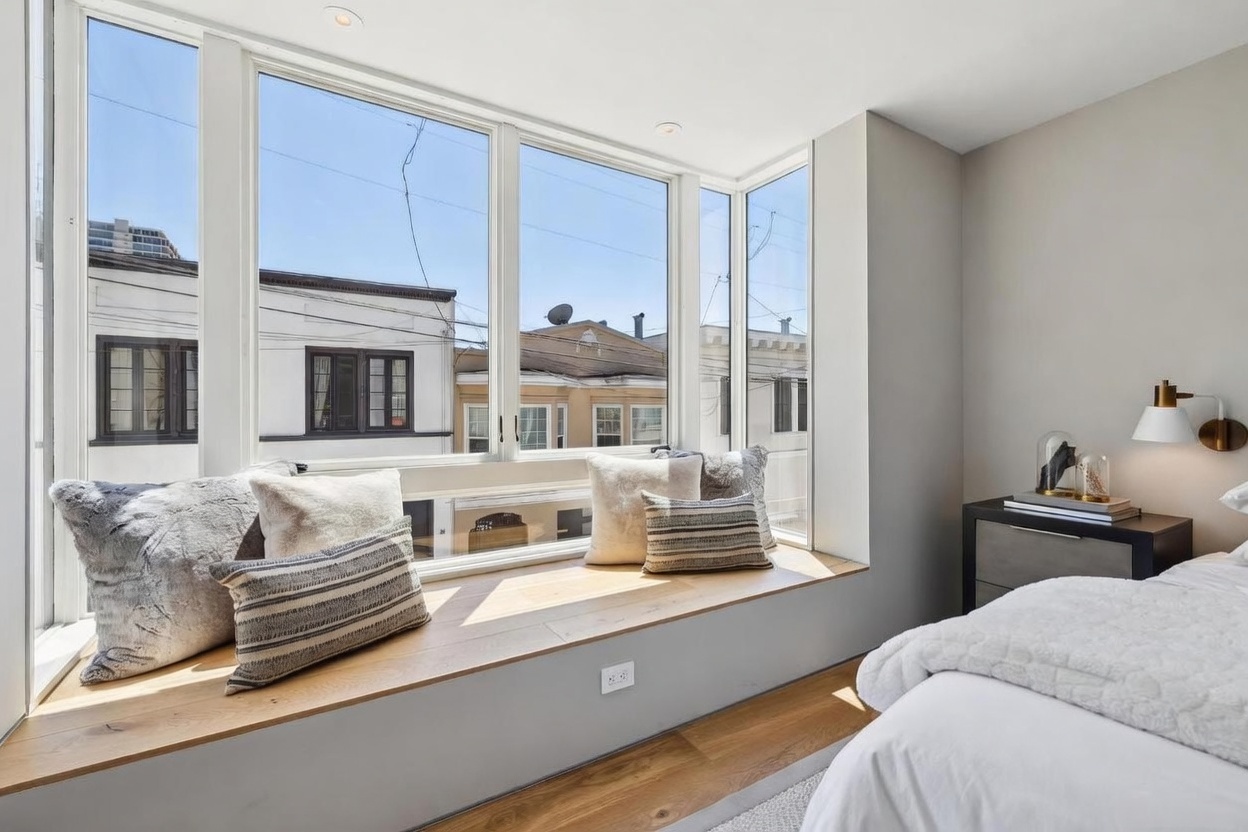 This bright bedroom features a prominent bay window with a built-in light wood bench seat, adorned with plush pillows and cushions. The space is bathed in natural light, highlighting the clean, neutral walls and the corner of a bed with a textured white coverlet. A modern bedside table with a sleek lamp sits to the right, creating a cozy and inviting atmosphere.