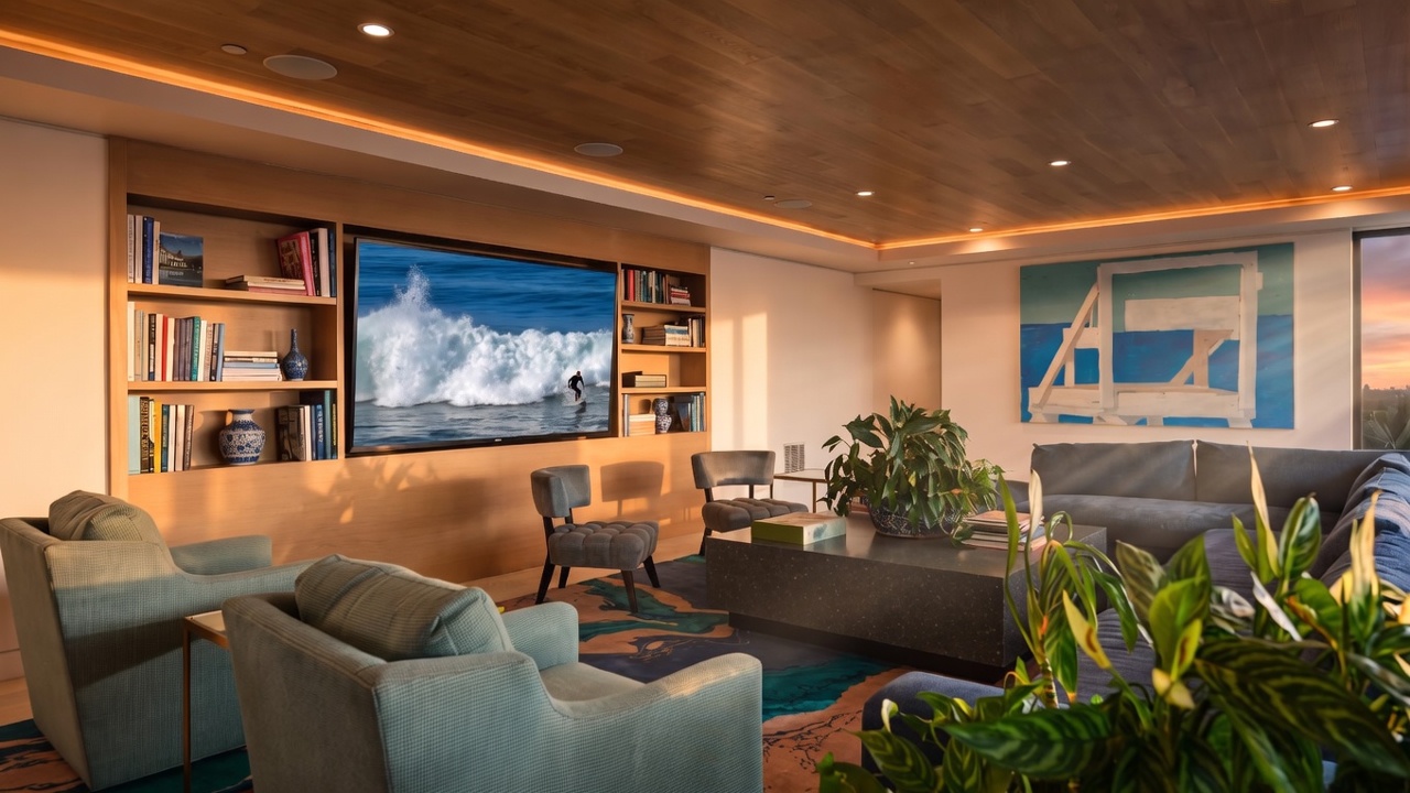 This contemporary living room features a warm, light wood-paneled ceiling with recessed lighting and a built-in media wall with integrated shelving. The space is furnished with plush teal armchairs, a dark gray sofa, and a large, low-profile stone coffee table, all anchored by a vibrant, abstract-patterned area rug. A large piece of modern art depicting a lifeguard tower hangs on the wall, contributing to the sophisticated, coastal-inspired aesthetic.