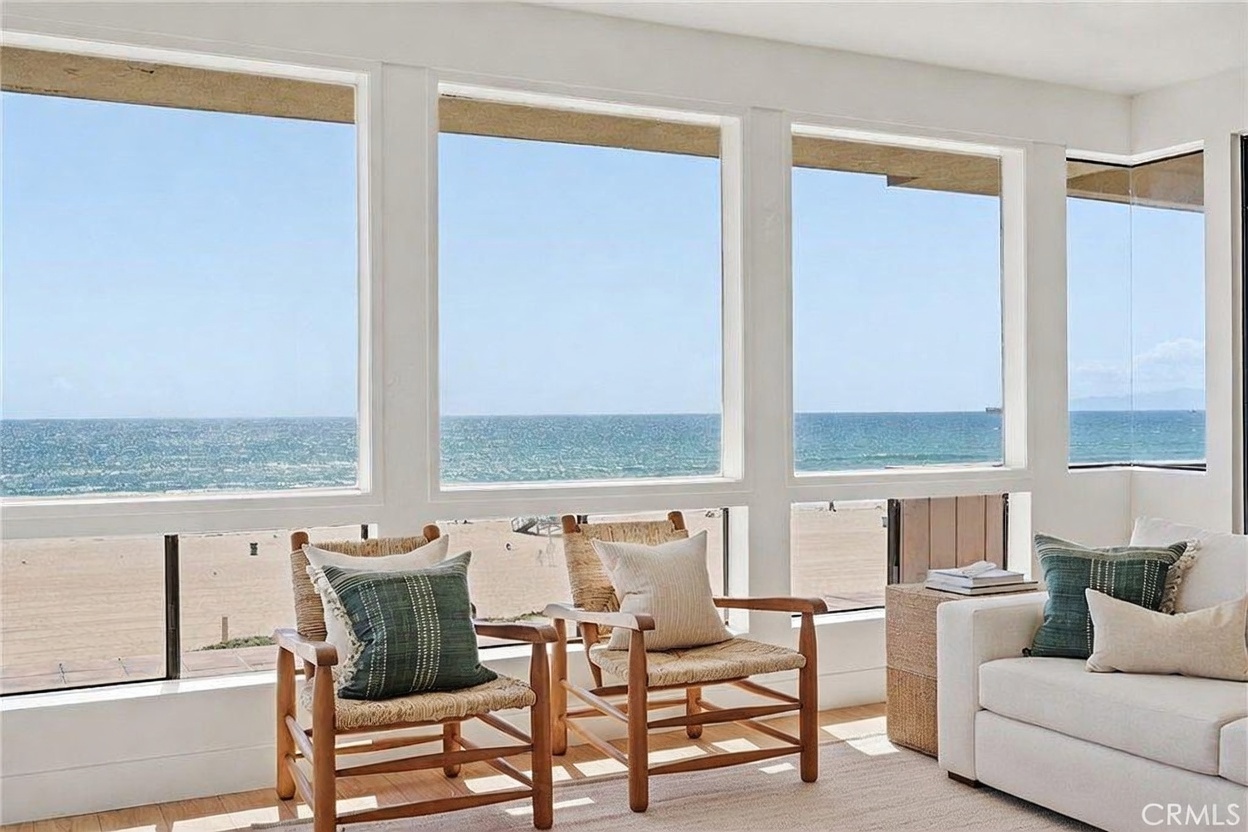 This bright living room features a wall of large windows that provide an expansive, unobstructed view of the ocean and sandy beach. The space is furnished with two light wood armchairs with patterned cushions and a white sofa, centered around a light-toned wooden coffee table. The overall aesthetic is airy and coastal, emphasizing the seamless connection between the indoor living area and the natural scenery outside.