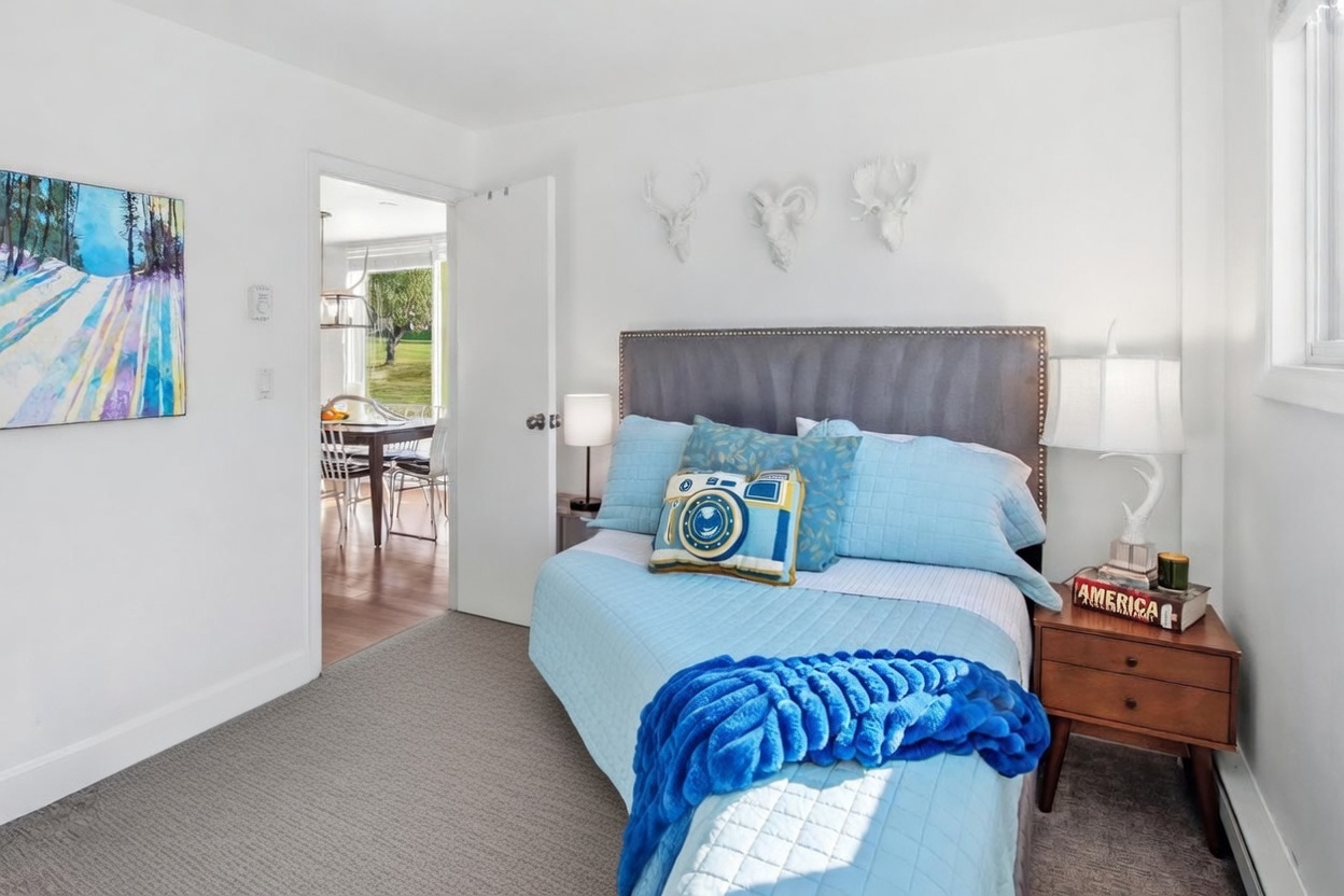 This cozy bedroom features a queen-sized bed with a tufted grey headboard, dressed in light blue quilted bedding and a vibrant blue textured throw blanket. The room is decorated with whimsical wall-mounted animal sculptures and a playful camera-shaped accent pillow, creating a cheerful and inviting atmosphere. A wooden nightstand with a lamp sits beside the bed, while an open doorway provides a glimpse into an adjacent dining area with a view of the outdoors.