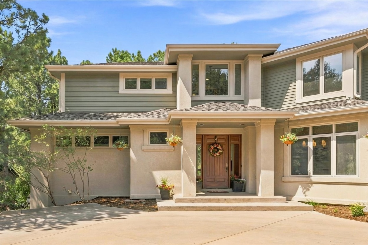 This front-facing view of a multi-level home showcases a blend of light-colored stucco and horizontal siding, creating a warm and inviting curb appeal. The entryway is framed by substantial columns and a covered porch, leading to a wooden front door adorned with a seasonal wreath. Large windows, including a prominent upper-level window bank, allow for natural light, while hanging planters and potted flowers add a touch of color to the concrete driveway and entrance area.