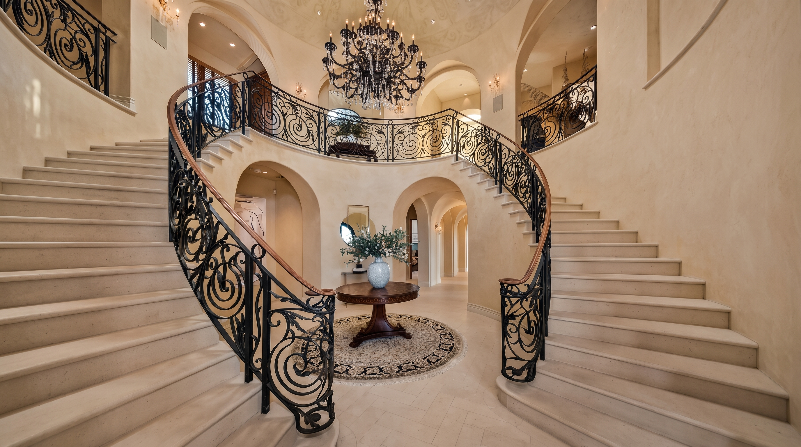 This grand foyer features a dramatic dual staircase with intricate wrought-iron railings that curve elegantly toward the upper level. A large, ornate chandelier serves as the centerpiece, hanging above a circular table placed on a patterned rug in the center of the light-toned stone floor. The space is defined by its high, arched doorways and neutral walls, creating an expansive and luxurious atmosphere.