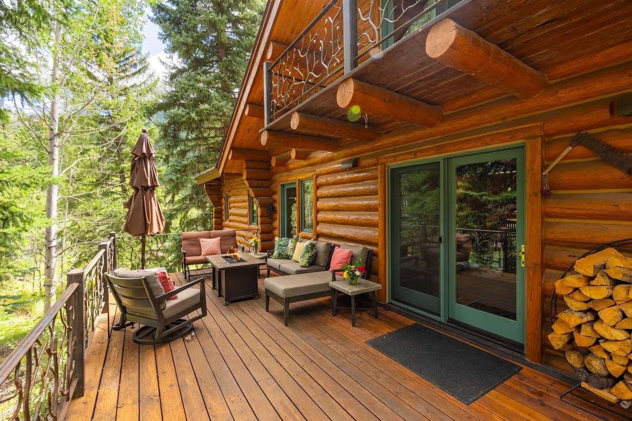 This inviting log cabin deck features a cozy outdoor seating area complete with a fire pit, plush sofas, and a rocking chair. The space is framed by rustic log walls and a decorative metal railing, with a stack of firewood neatly stored near the green-framed glass doors. The perspective captures a serene, elevated outdoor living space nestled among lush trees, perfect for relaxing in a natural setting.