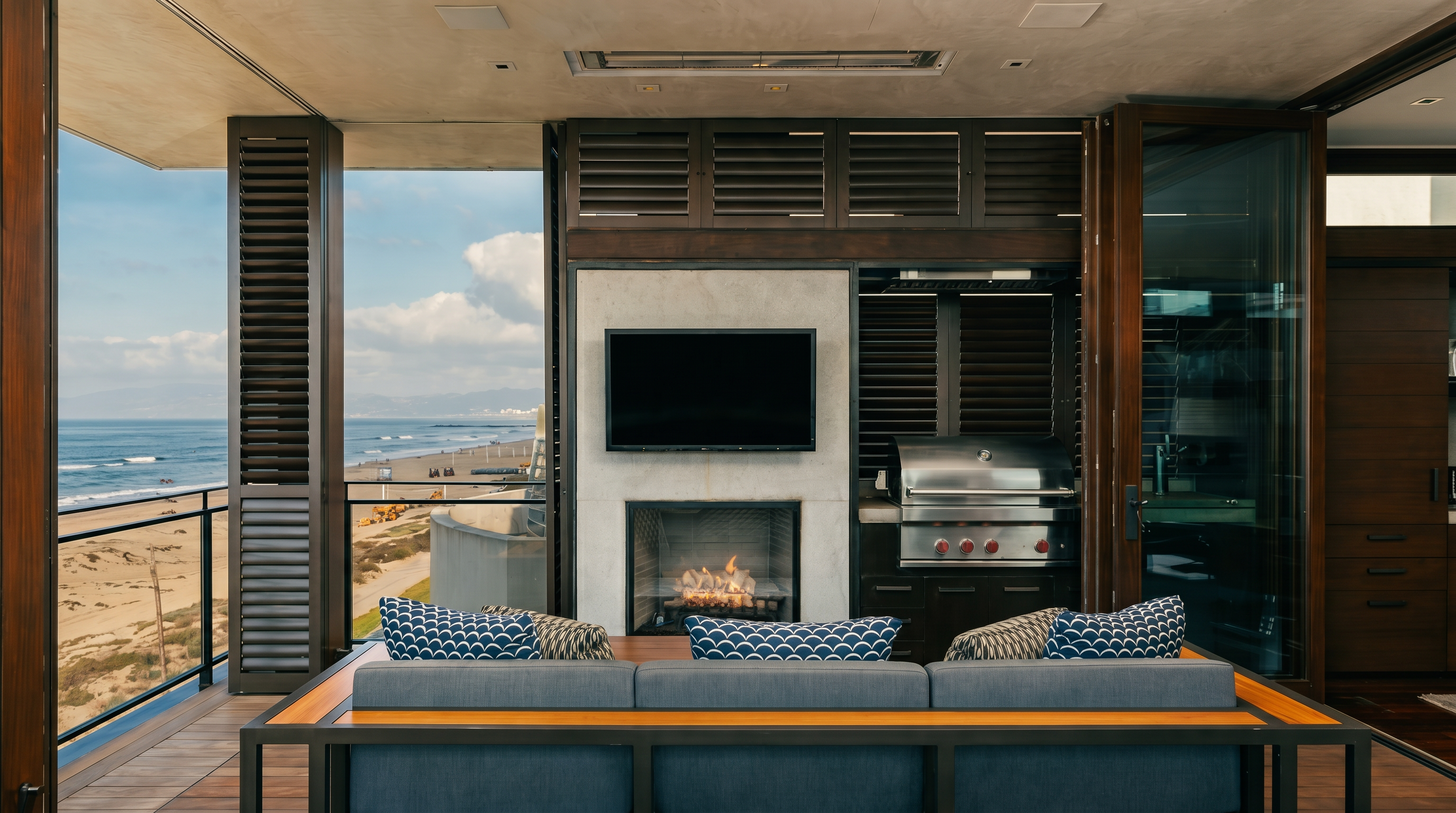 This luxurious outdoor living space features a covered balcony overlooking the ocean, complete with a built-in grill and a central fireplace set into a stone-clad chimney breast. A large, modern sofa with patterned cushions faces the fireplace, creating an inviting lounge area. The space is framed by dark wood-toned shutters and opens directly to a scenic coastal view, blending indoor comfort with an expansive outdoor atmosphere.