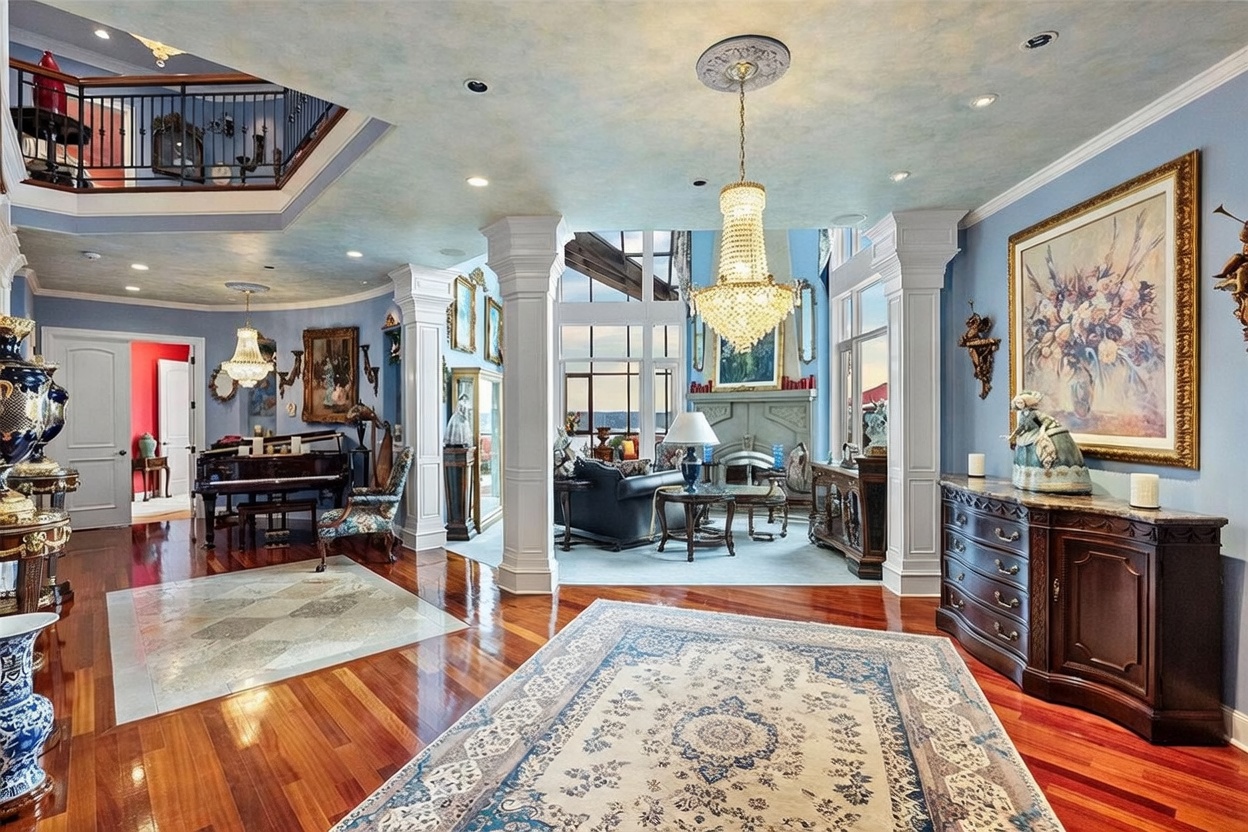 This elegant dining room features a formal dark wood table set beneath a grand crystal chandelier, creating a sophisticated atmosphere. The space is adorned with ornate decor, including a large framed floral painting, classical columns, and polished hardwood floors that transition into a decorative stone inlay. A view into the adjacent living area reveals tall windows that allow natural light to fill the open-concept floor plan.