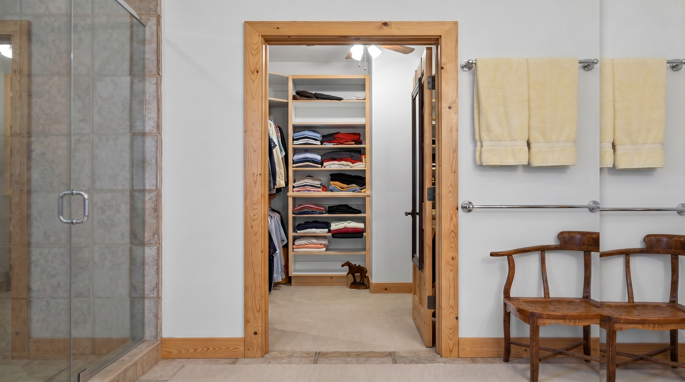 This interior view showcases a well-organized walk-in closet accessed through a rustic wood-trimmed doorway from a bathroom. The closet features open shelving stacked neatly with folded clothing, hanging rods for shirts, and a carpeted floor. Outside the closet, a simple wooden chair and towel racks on the wall suggest a functional and cozy bathroom layout.