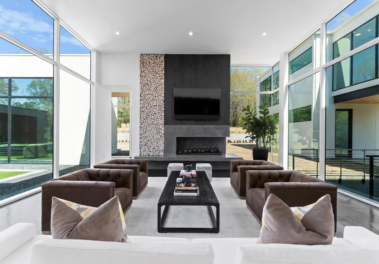 This expansive living room features a striking central fireplace with a dark, textured surround and an adjacent decorative wall of stacked wood rounds. The space is framed by floor-to-ceiling windows on both sides, offering abundant natural light and views of the surrounding landscape. Four brown leather tufted armchairs are arranged around a dark rectangular coffee table, creating a sophisticated and symmetrical seating area.