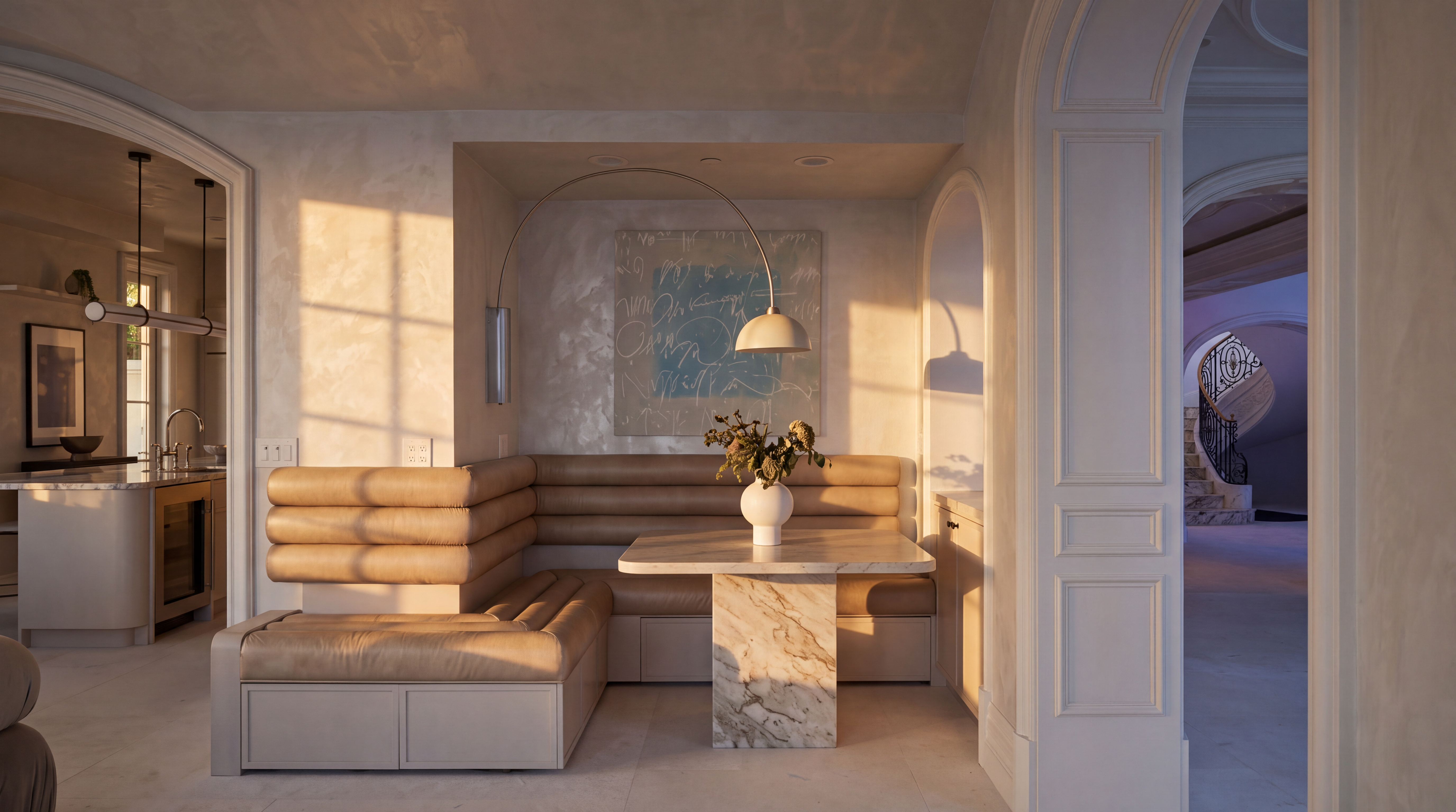 This elegant breakfast nook features a custom-built, white upholstered banquette set into a recessed alcove, illuminated by integrated under-seat lighting and a sleek, arched floor lamp. A central table with a light-toned top and a dark, solid base sits in front of a piece of abstract wall art, while an arched doorway leads to a glimpse of a grand staircase in the background. The space is finished in a monochromatic, light-toned palette that emphasizes a clean, sophisticated, and contemporary aesthetic.