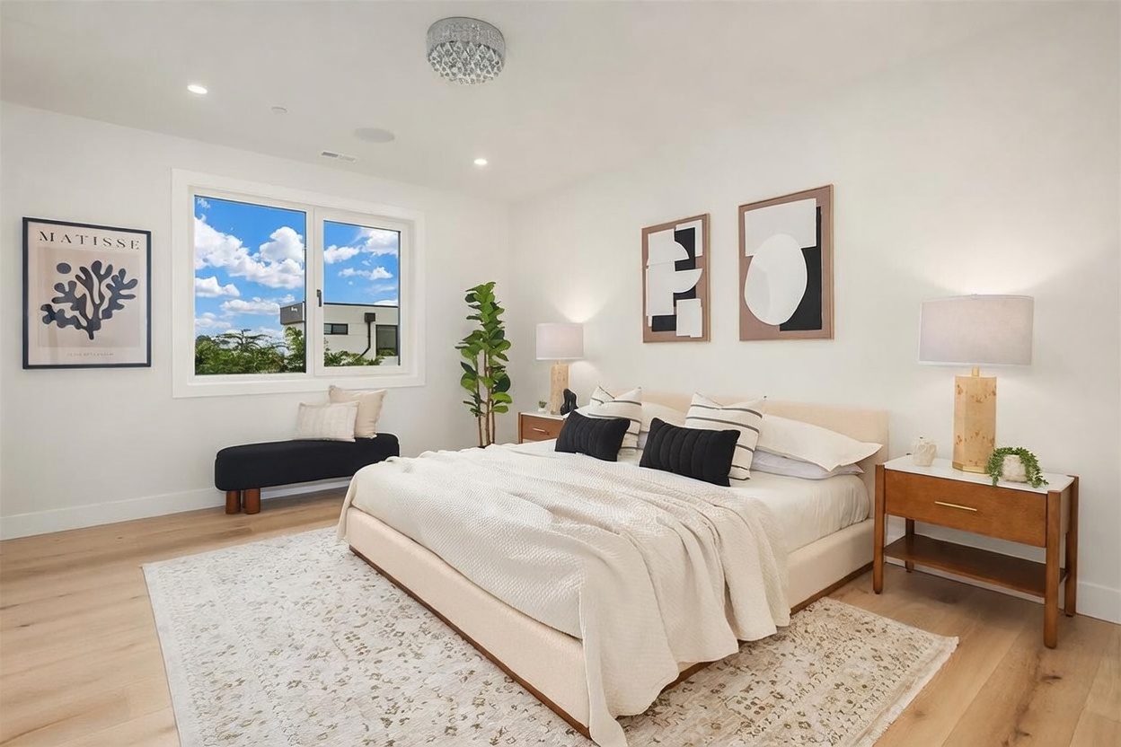 This bright and airy primary bedroom features a neutral color palette with light wood flooring and a large area rug. The room is anchored by a plush bed with layered pillows and a textured throw, flanked by two wooden nightstands with matching lamps. A Matisse print and abstract wall art add a touch of modern sophistication, while a window bench provides a cozy seating area overlooking the exterior.