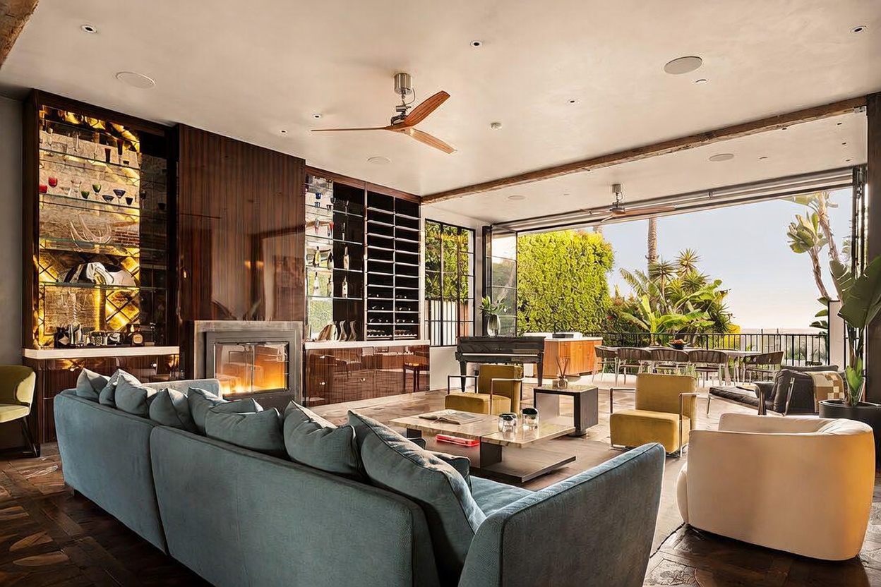 This expansive, open-concept living room features a sophisticated design with a large, plush sofa facing a fireplace set into a dark wood-paneled wall. The space seamlessly transitions to an outdoor patio through massive, retractable glass walls, creating an indoor-outdoor living experience. A grand piano and stylish seating area add to the luxurious atmosphere, while the warm lighting and polished floors enhance the overall sense of elegance.