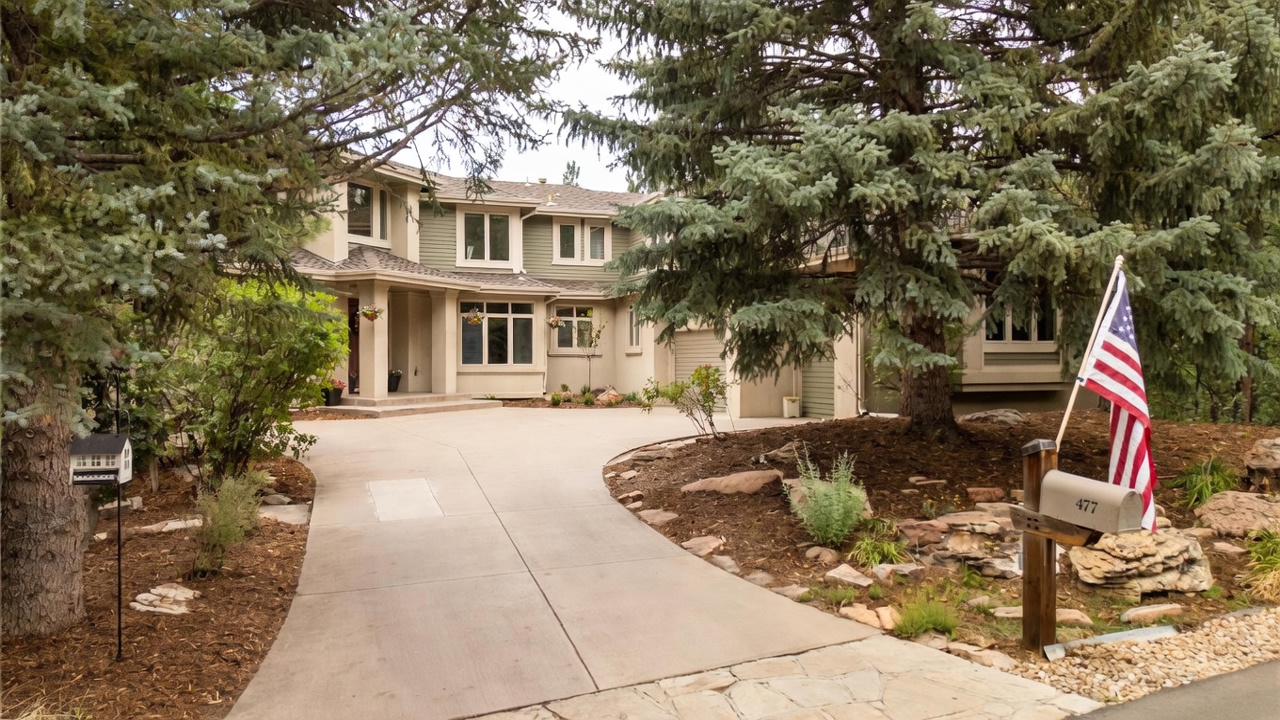 This two-story suburban home features a blend of light-colored siding and stone accents, nestled among mature evergreen trees. A curved concrete driveway leads to the front entrance and a multi-car garage, while a mailbox with an American flag stands prominently in the foreground. The landscaping includes natural rock features and established shrubs, creating a private and wooded atmosphere.
