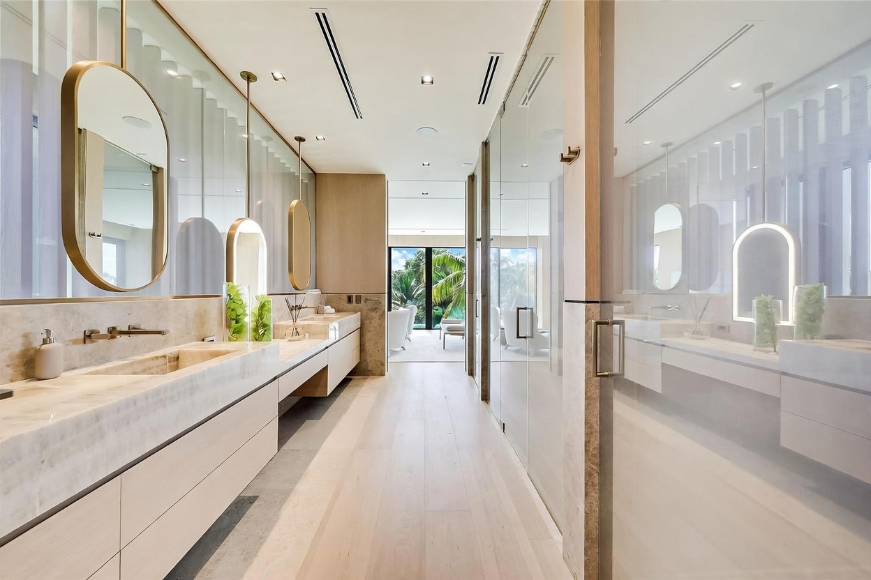This luxurious primary bathroom features a long, dual-vanity setup with stone countertops and integrated sinks, complemented by elegant, suspended oval mirrors. The space is characterized by a clean, modern aesthetic with light wood flooring and glass-enclosed shower or water closet areas. A central hallway leads the eye toward a bright, open living space with large glass doors that provide a view of lush greenery outside.
