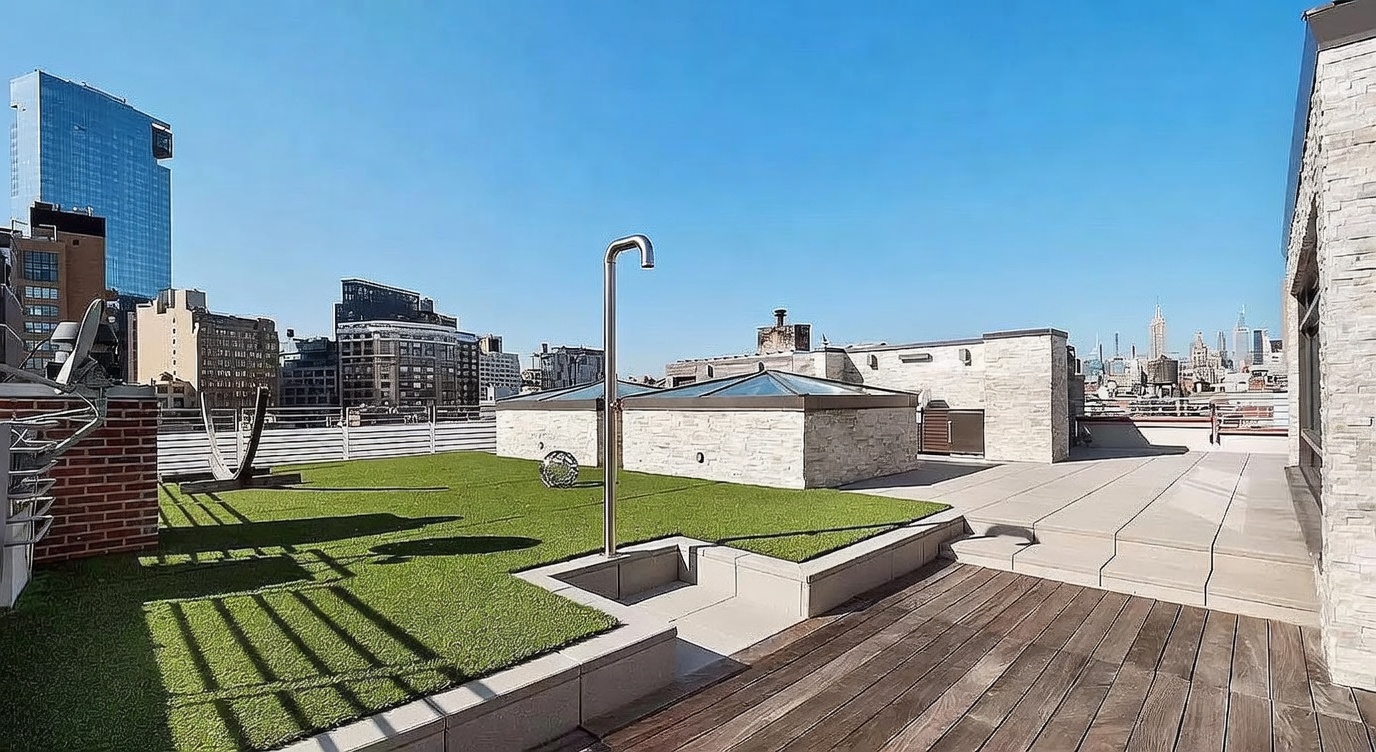 This expansive rooftop terrace offers a sophisticated outdoor living space with a blend of artificial turf, wood decking, and stone-tiled areas. A modern outdoor shower stands prominently on the lawn, while stone-clad structures provide architectural interest and privacy. The setting provides a panoramic view of the city skyline, creating an impressive urban retreat.