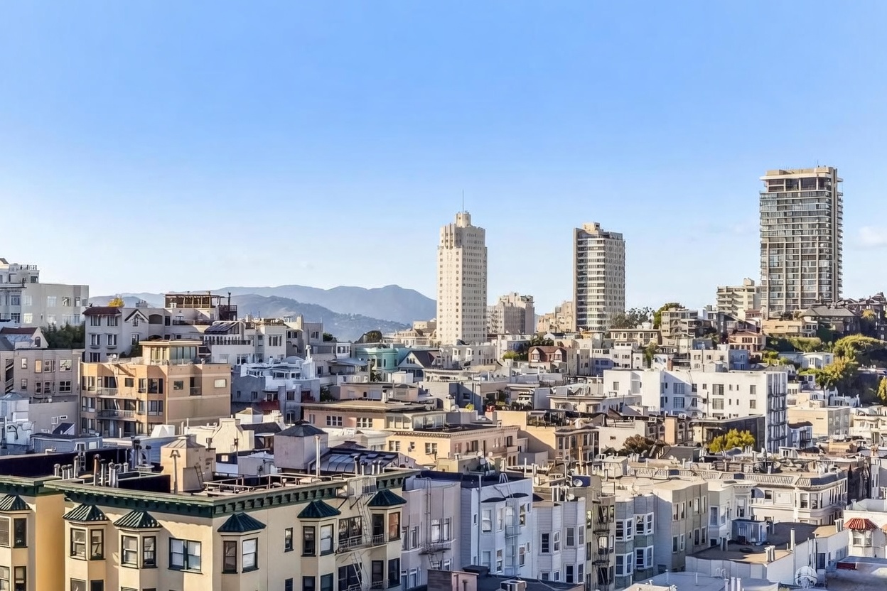 This elevated, wide-angle shot captures a dense urban cityscape, showcasing a mix of historic and modern residential architecture. The foreground features classic multi-story buildings with bay windows, while the background reveals prominent high-rise towers set against a backdrop of rolling hills under a clear blue sky. The perspective provides a comprehensive view of the neighborhood's density and its integration with the surrounding natural landscape.