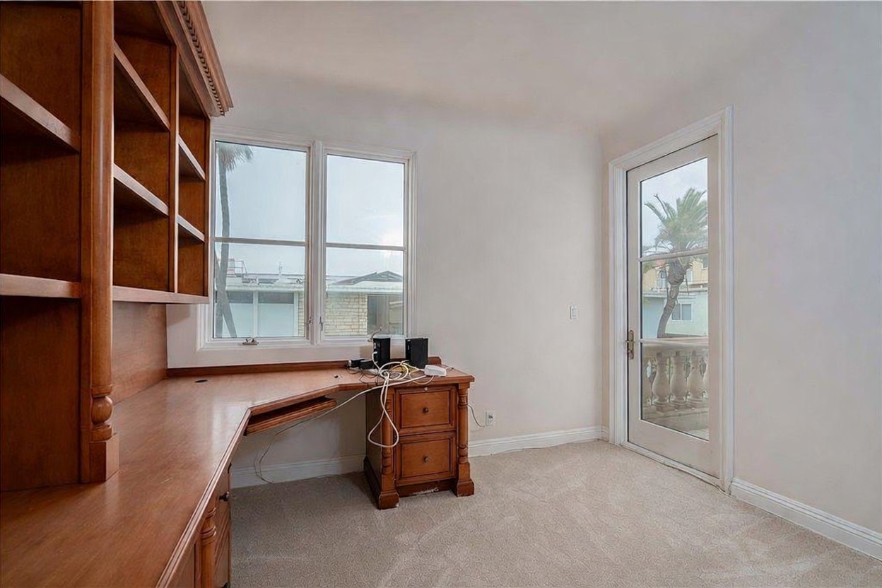 This home office features a built-in wooden desk unit with integrated shelving and a matching cabinet, providing ample workspace and storage. A large window allows natural light to fill the room, while a glass-paned door provides direct access to an outdoor balcony. The space is finished with neutral carpeting and light-colored walls, creating a functional and quiet environment.