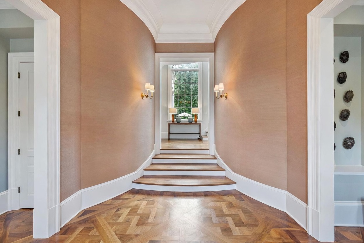 This elegant hallway features a barrel-vaulted ceiling and intricate patterned wood flooring that leads to a set of steps. The walls are finished in a textured, warm-toned wallcovering, accented by wall sconces and decorative urn-shaped sculptures on either side of the entryway. A distant view through a large window at the end of the hall provides natural light and a glimpse of the outdoors.