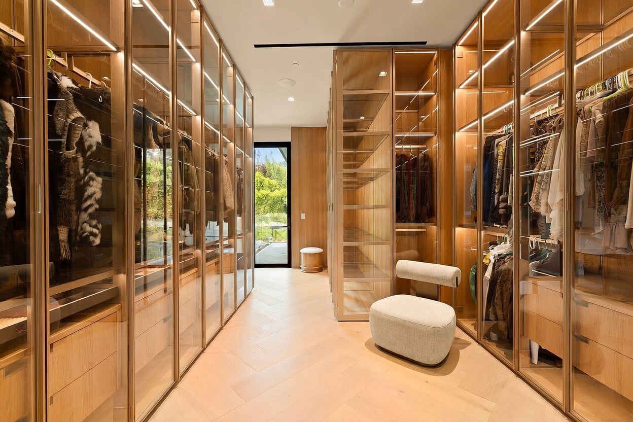 This luxurious walk-in closet features extensive light wood cabinetry with glass-front doors and integrated LED lighting, creating a sophisticated and organized display. A central, modern upholstered chair sits on the light-toned herringbone wood flooring, while a doorway at the end of the hall provides a view of the lush greenery outside. The space is designed with a clean, contemporary aesthetic that emphasizes both storage capacity and high-end style.