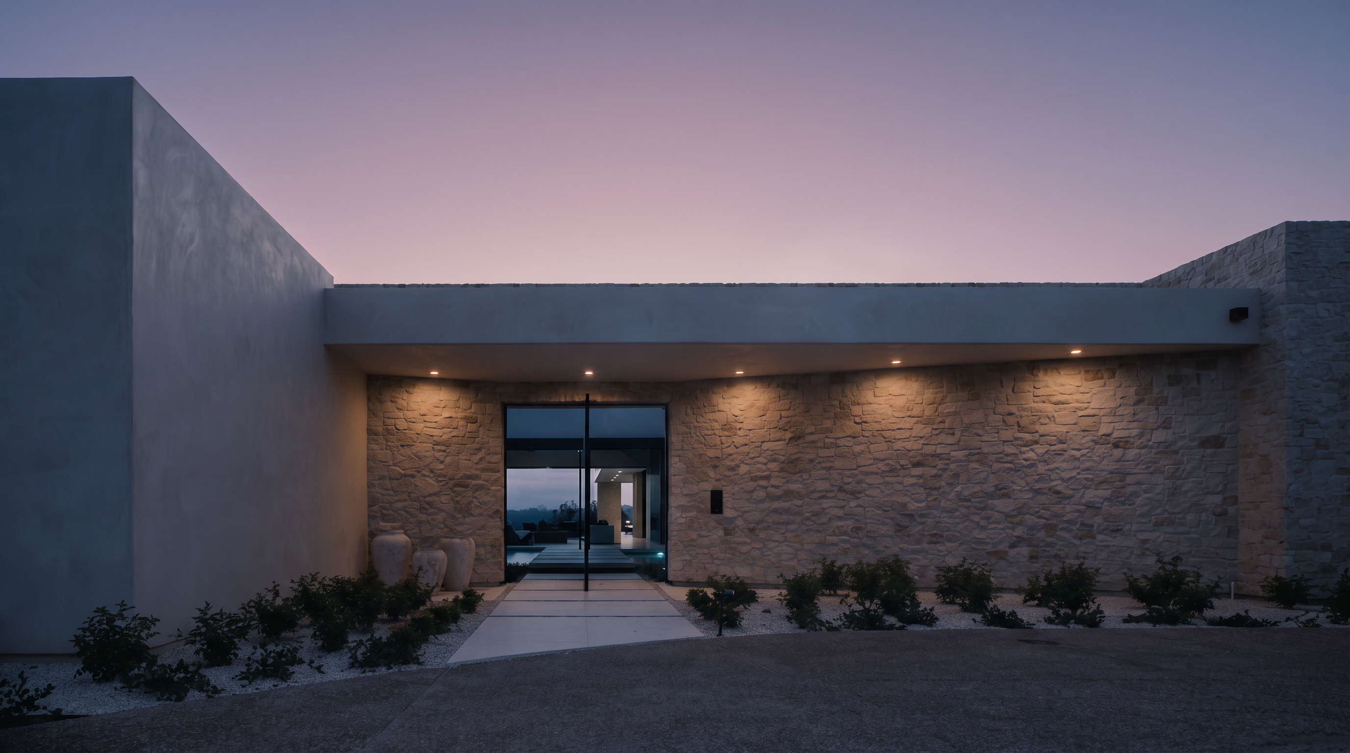 This striking modern entryway features a wide, flat roofline extending over a stone-clad facade, creating a dramatic and welcoming approach. A central glass door provides a clear view through the home to the rear, while recessed lighting illuminates the textured stone wall and the paved walkway. The scene is captured at dusk, with a soft, pastel-toned sky providing a serene backdrop to the clean, architectural lines of the residence.