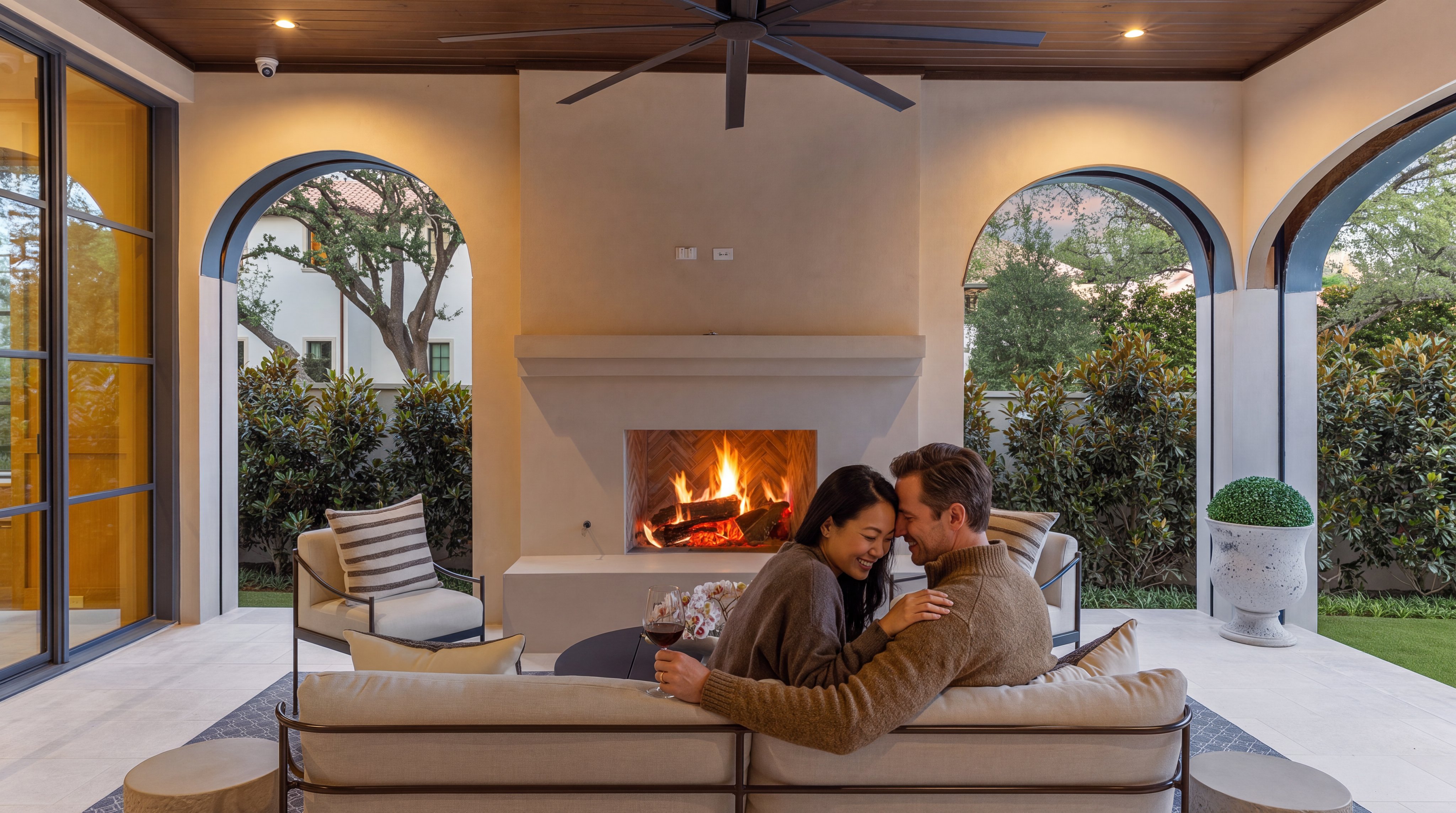 This luxurious covered outdoor patio features a central fireplace with elegant arched openings, providing an intimate and sophisticated space for relaxation. The space includes a comfortable seating arrangement with stylish, earth-toned furniture, perfect for hosting or unwinding. With its high-end finishings, built-in ceiling fan, and framed views of the landscaped backyard, this area exemplifies upscale outdoor entertaining.