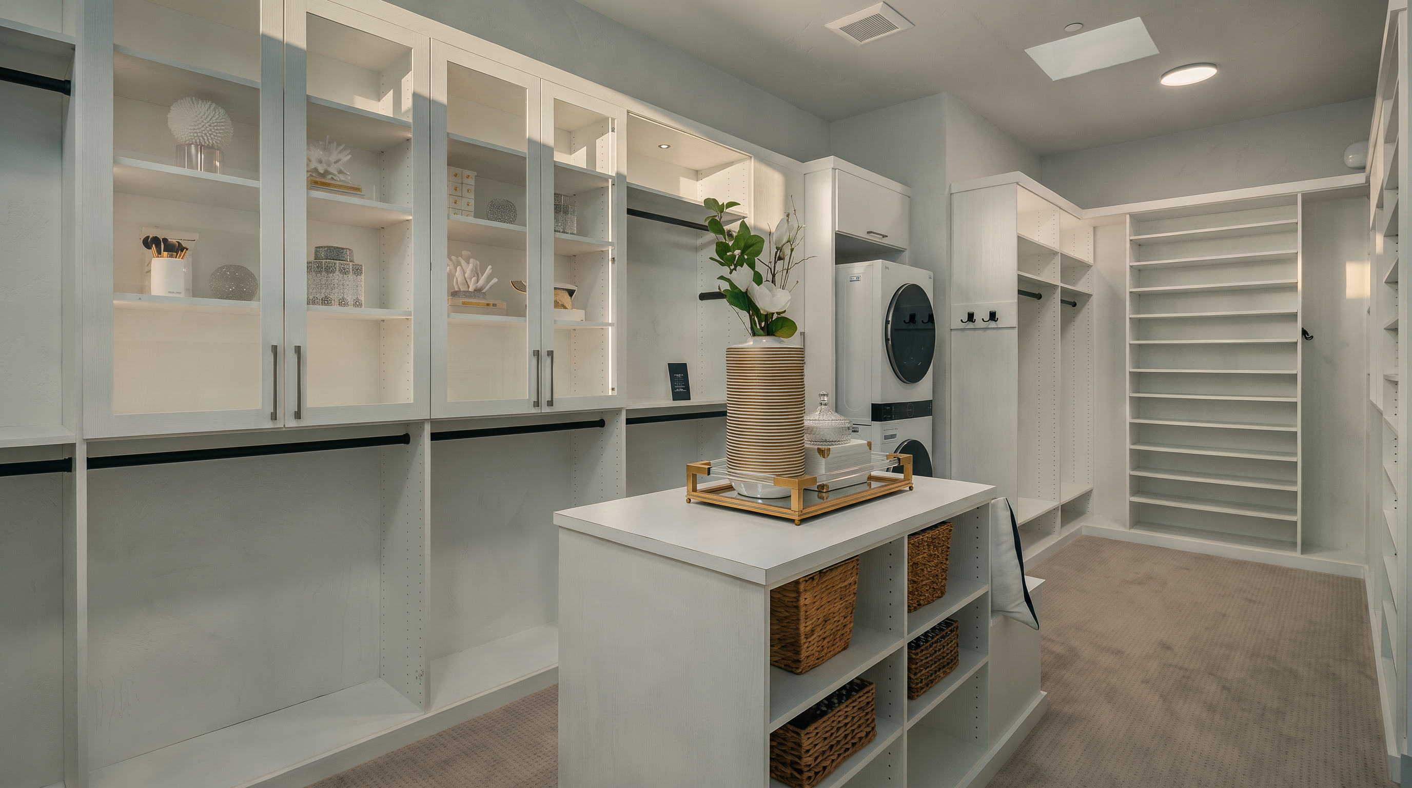 This spacious, high-end walk-in closet features extensive white custom cabinetry with glass-fronted upper storage and integrated hanging rods. A central island provides additional storage with open shelving and woven baskets, topped with a decorative vase and tray. The room is finished with neutral carpeting and includes a stacked washer and dryer unit, blending functionality with organized luxury.