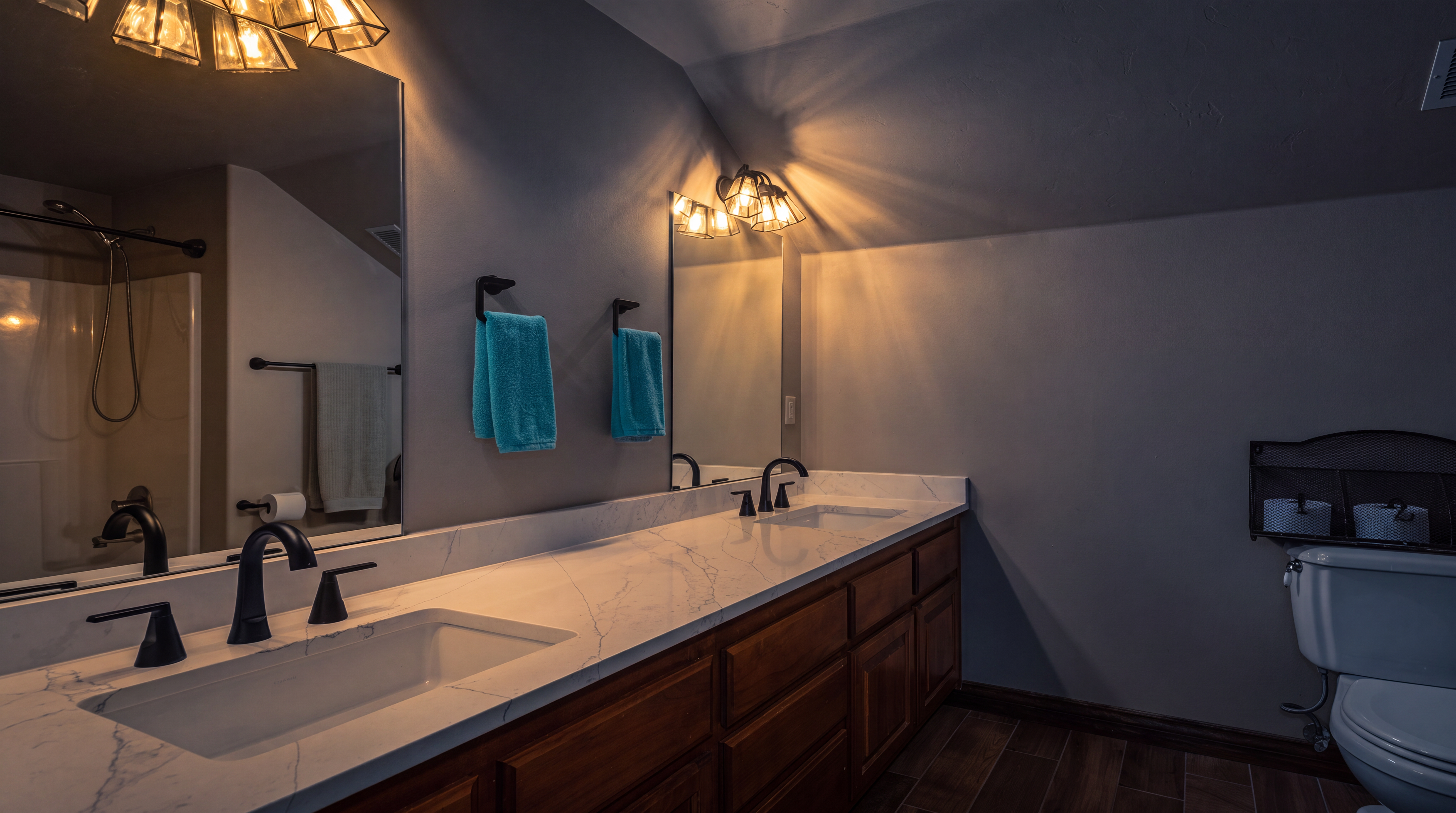 This functional bathroom features a long dual-sink vanity with warm-toned wooden cabinetry and a contemporary white marble-veined countertop. The space includes dark matte fixtures, dual mirrors with matching light sconces, and a shower-tub combination, all grounded by wood-look tile flooring. The neutral wall color creates a clean and welcoming atmosphere suitable for a guest or shared bathroom.