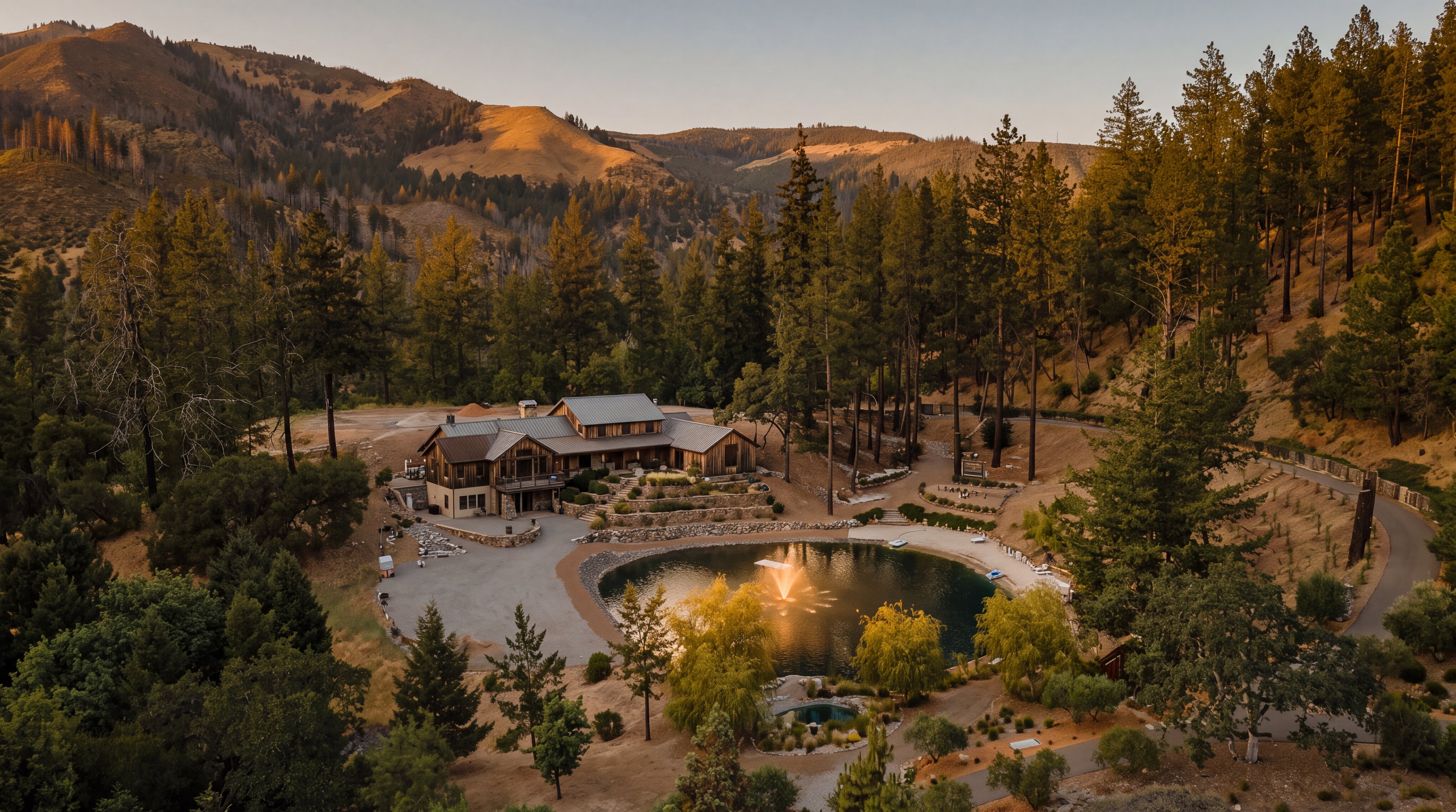 This high-angle aerial view captures a serene, expansive property featuring a large, circular pond with a central fountain as its focal point. The landscape is characterized by a mix of dry, golden-hued terrain and dense, mature evergreen trees, set against a backdrop of rolling hills under a clear blue sky. A paved path winds through the lower portion of the grounds, highlighting the natural beauty and secluded atmosphere of this rural estate.